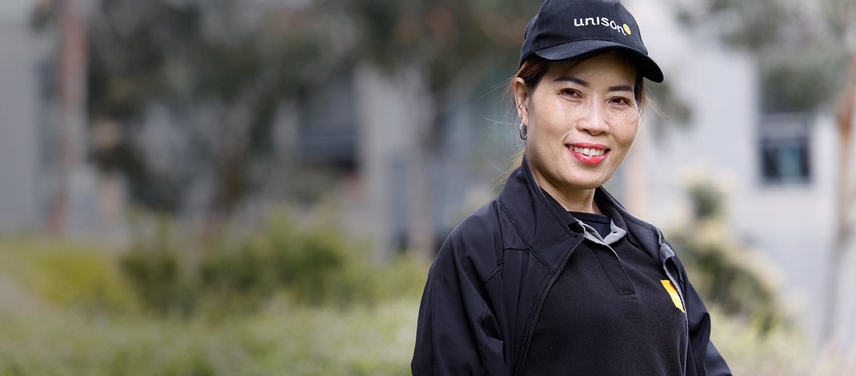 A woman wearing a black unison cap and black uniform stands outdoors, smiling at the camera with greenery and buildings blurred in the background.