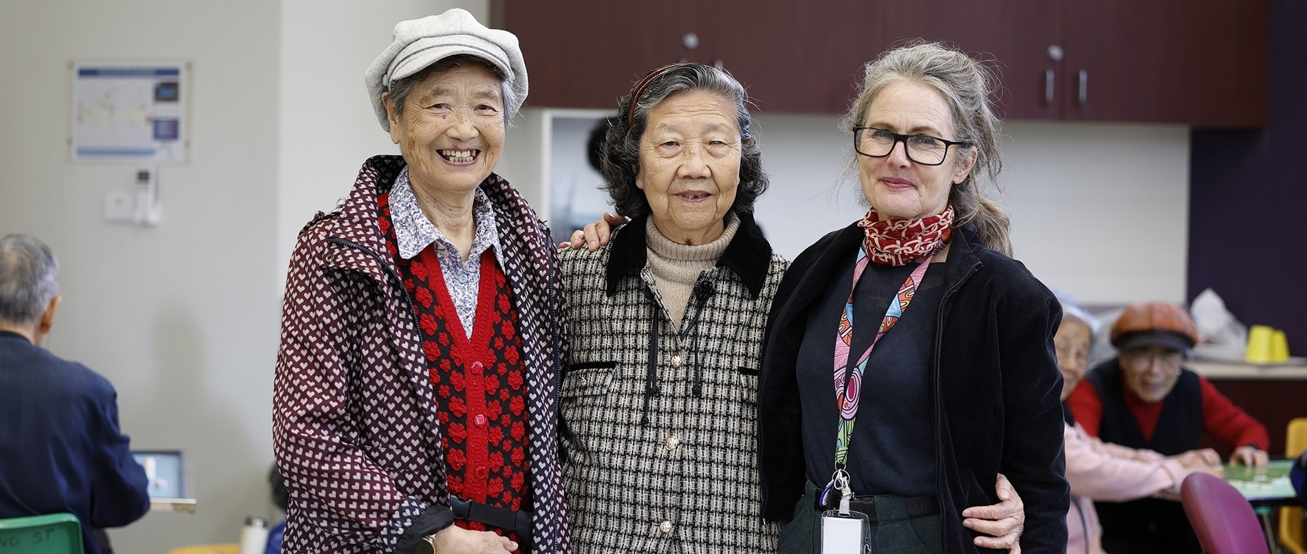 Three older women stand together indoors, smiling at the camera. Two wear patterned sweaters and hats; one wears glasses and a staff lanyard. Other people are visible in the background, sitting at tables.
