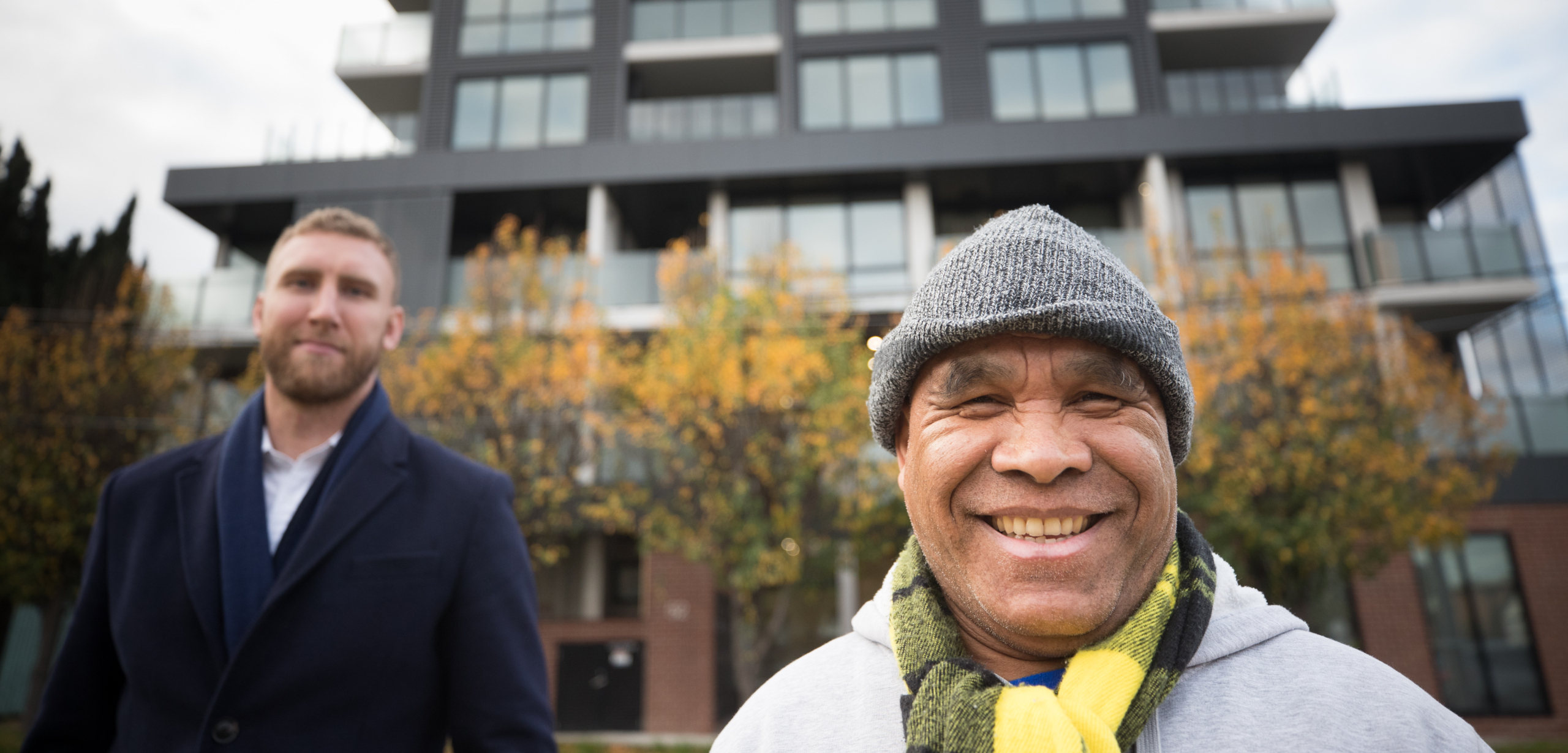 Two men stand outside a modern apartment building. The man in front is smiling, wearing a gray beanie and a striped scarf; the man in the background wears a dark coat. Trees with autumn leaves are behind them.