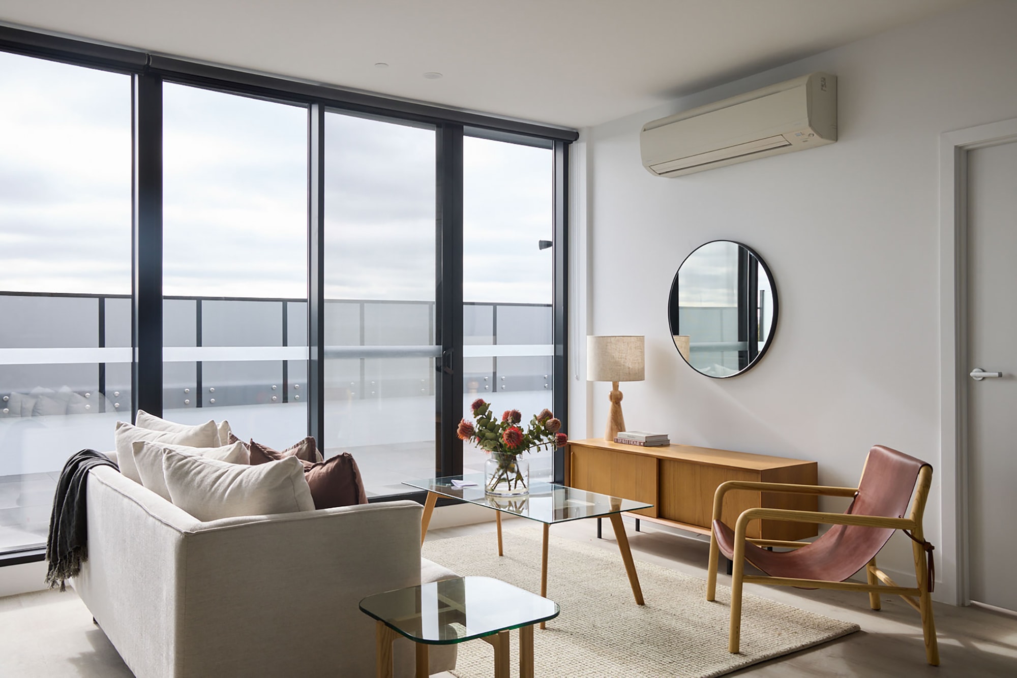 A modern living room with floor-to-ceiling windows, a beige sofa, a glass coffee table, a wooden sideboard with a lamp and flowers, a round wall mirror, a pink chair, and an air conditioner above the door.