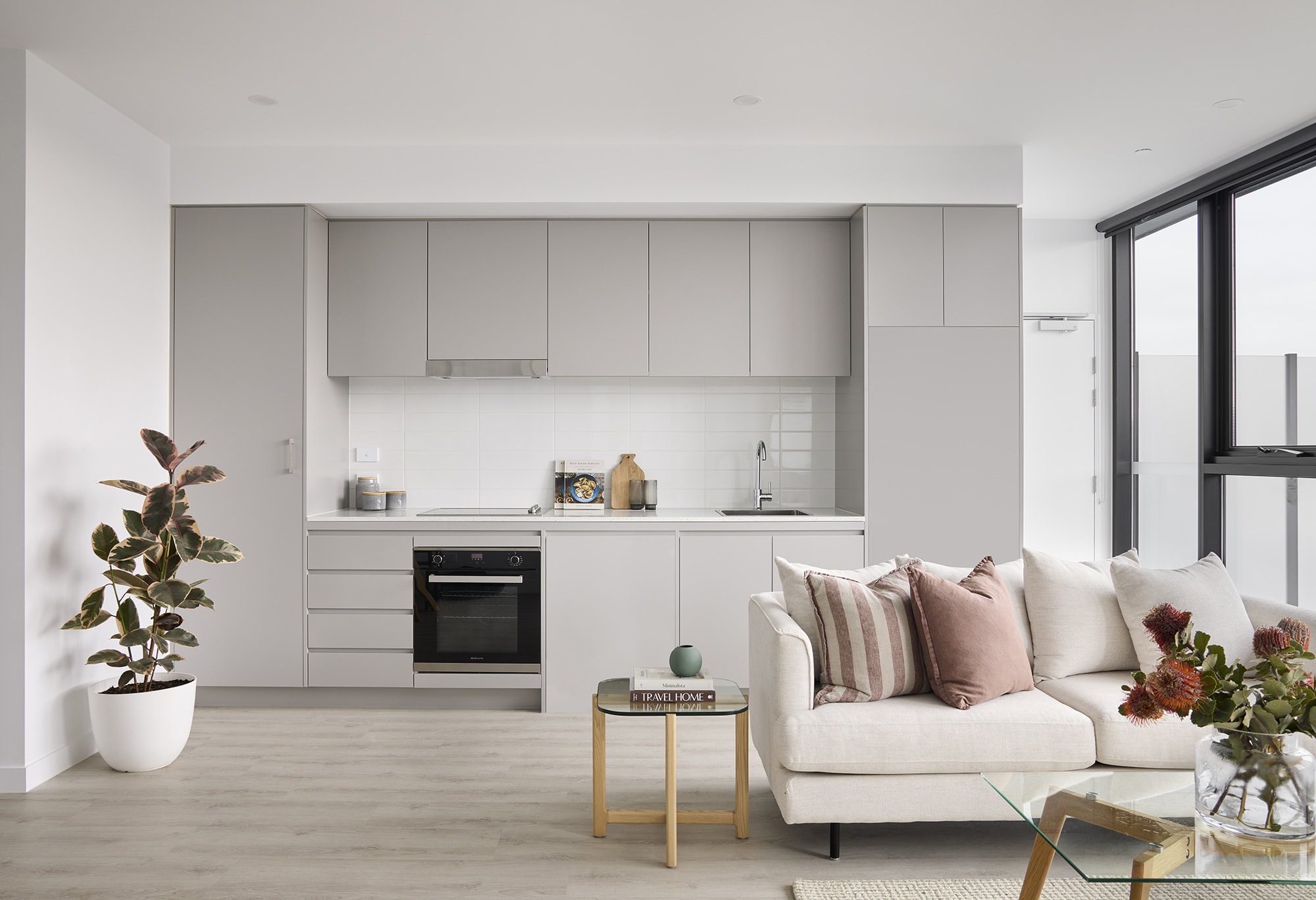 Modern, minimalist kitchen and living area with light grey cabinets, built-in oven, and white backsplash. A potted plant, beige sofa with pink cushions, and a small coffee table with books complete the space. Large windows provide natural light.