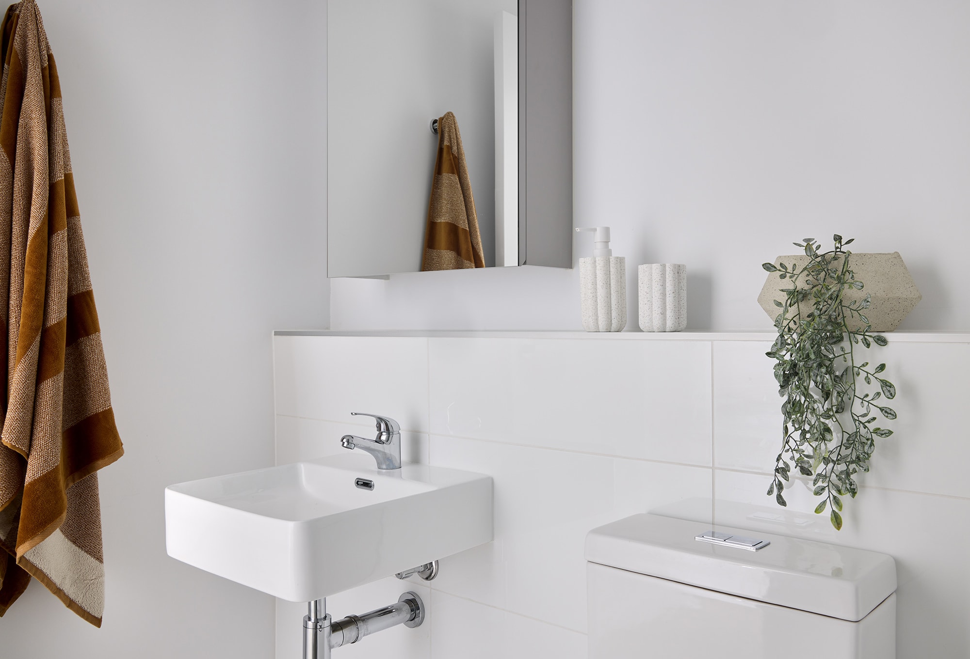 A modern bathroom with a white sink, toilet, and wall tiles. A brown striped towel hangs on the wall, and a decorative vase with greenery and textured jars sit on a shelf below a mirror.