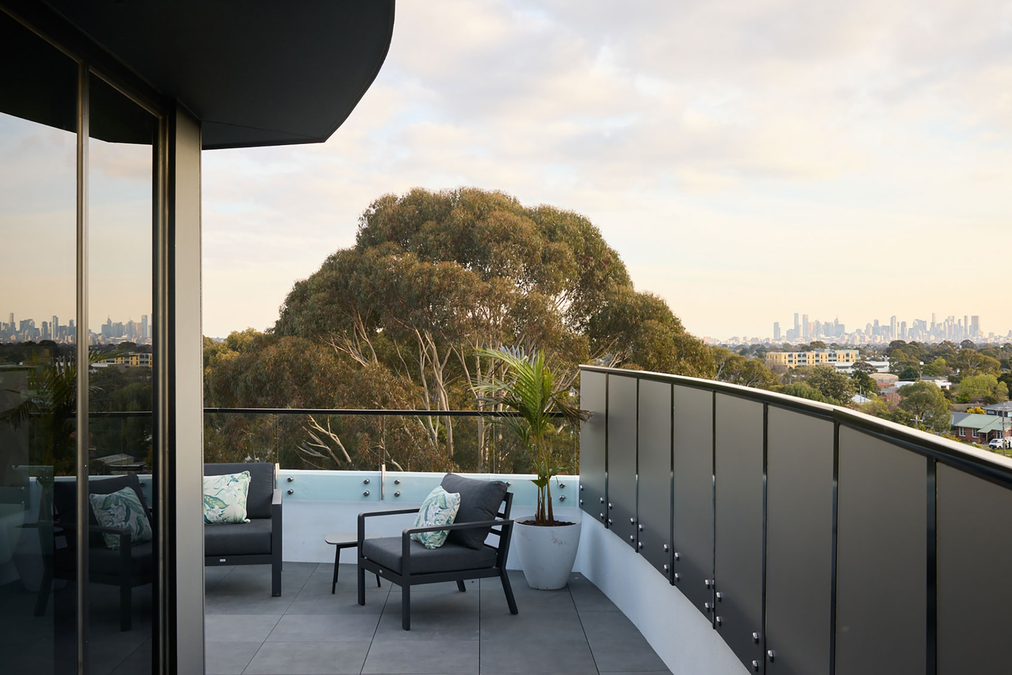 A modern balcony with glass railing, two chairs, a small table, and a potted plant overlooks trees and a distant city skyline under a partly cloudy sky.