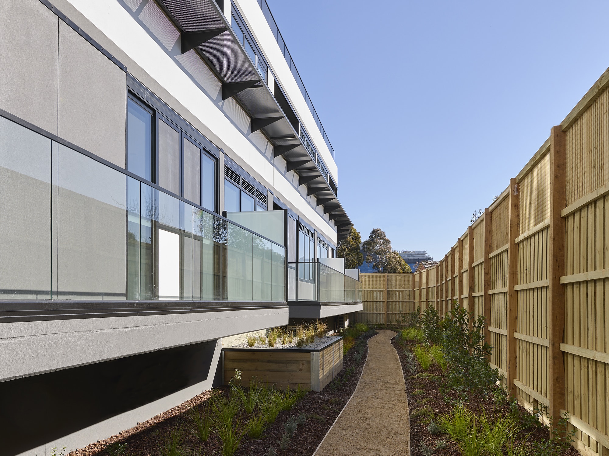 A modern building with glass balconies overlooks a narrow, curved gravel path bordered by plants and a tall wooden fence under a clear blue sky.