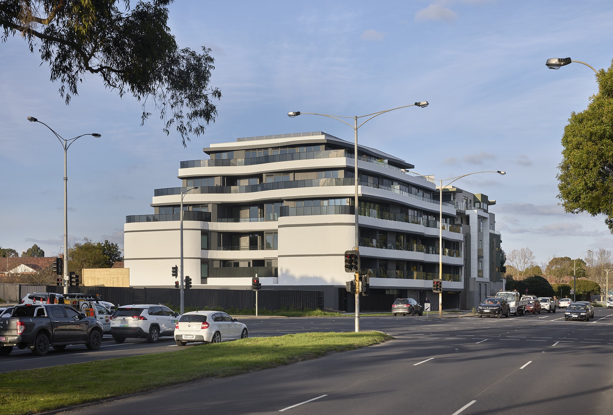 A modern, multi-story apartment building with curved balconies sits at a busy intersection with several cars and traffic lights, under a partly cloudy sky. Trees and suburban houses are visible in the background.