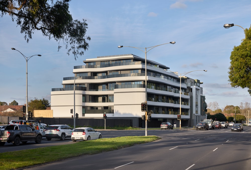 A modern, multi-story apartment building with glass balconies stands beside a busy intersection with cars and traffic lights under a partly cloudy sky. Trees and houses are visible in the background.