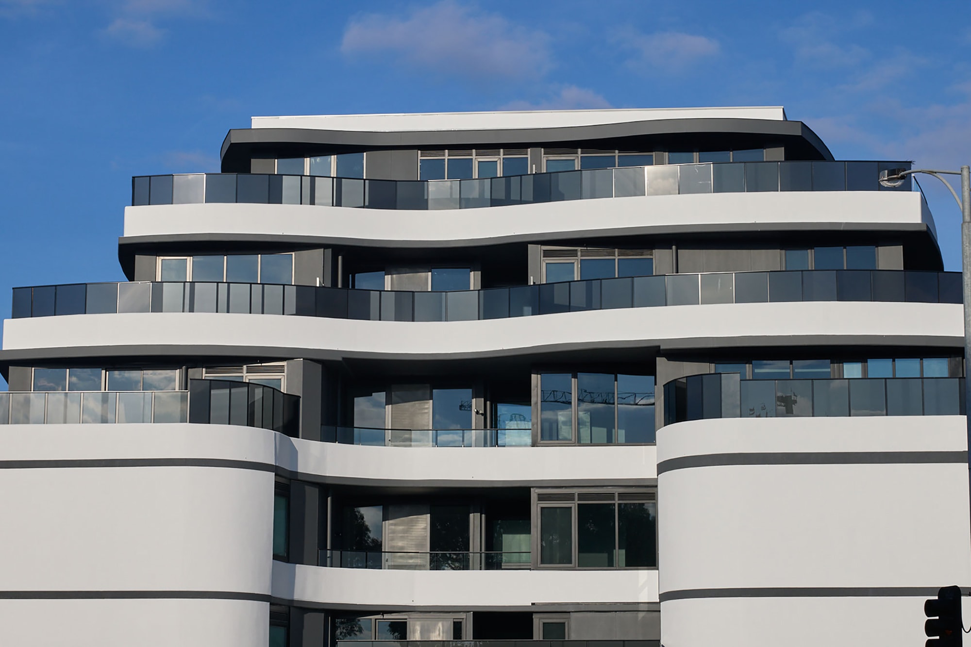 A modern, multi-story building with curved white and dark balconies and glass railings, set against a blue sky with scattered clouds.
