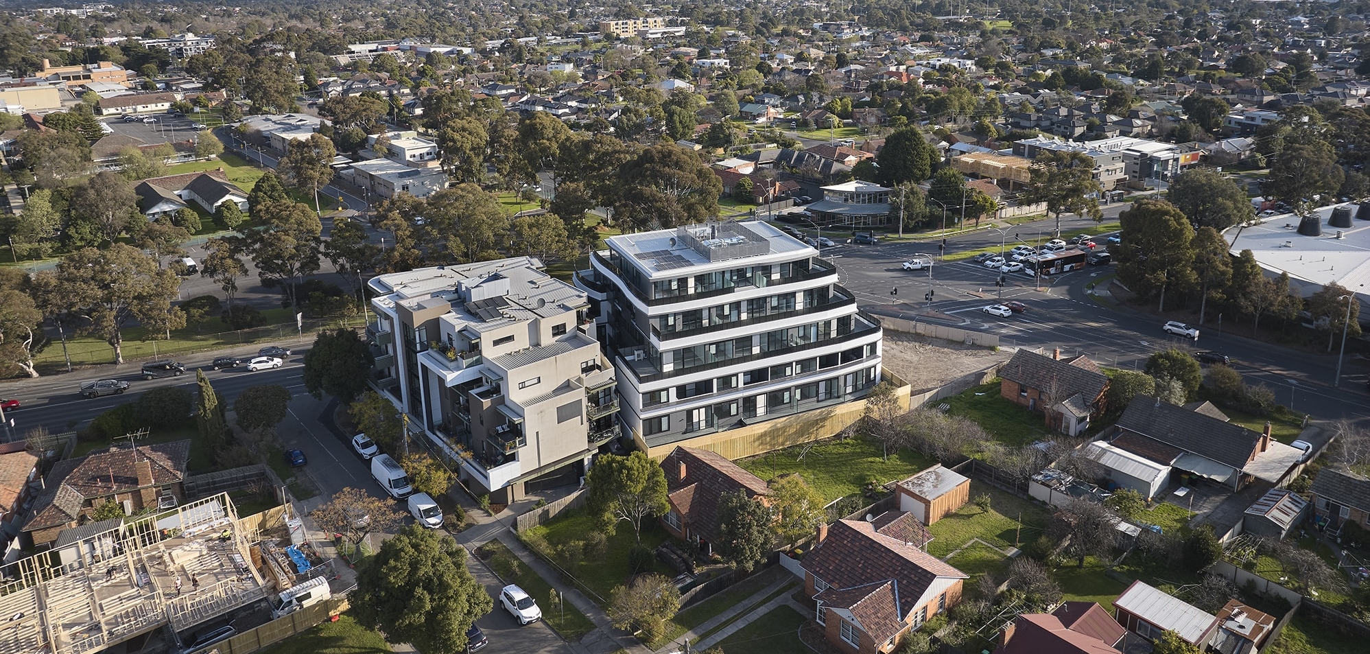 Aerial view of a modern multi-story apartment building in a suburban neighborhood, surrounded by houses, trees, roads, and intersections on a clear day.