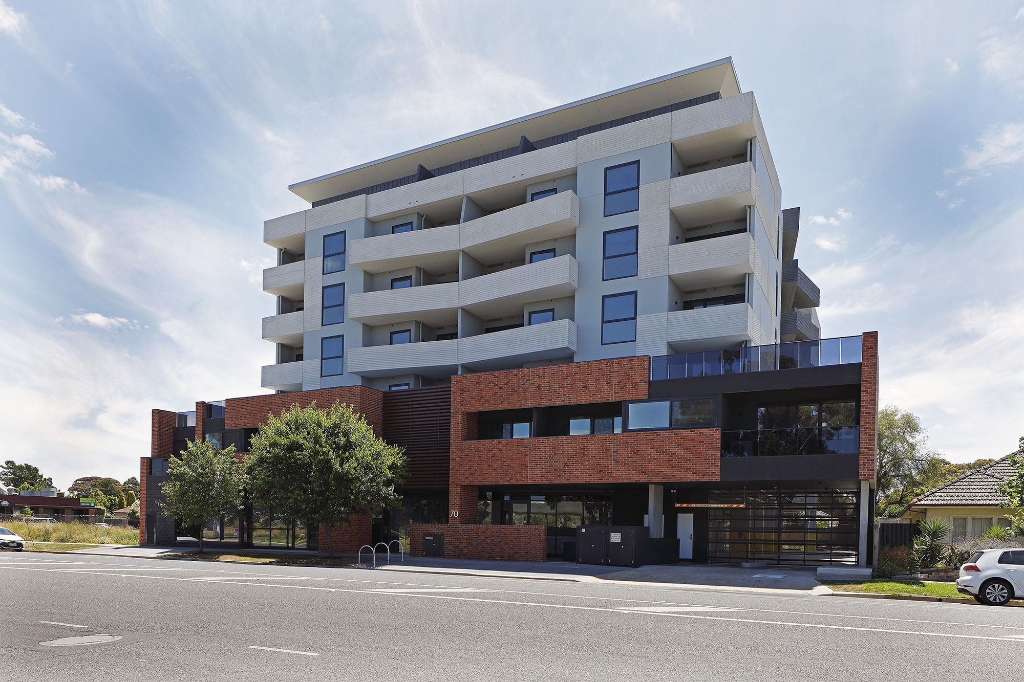 A modern multi-story apartment building with brick and light-colored facades, featuring balconies, large windows, and trees in front, situated along a quiet urban street.