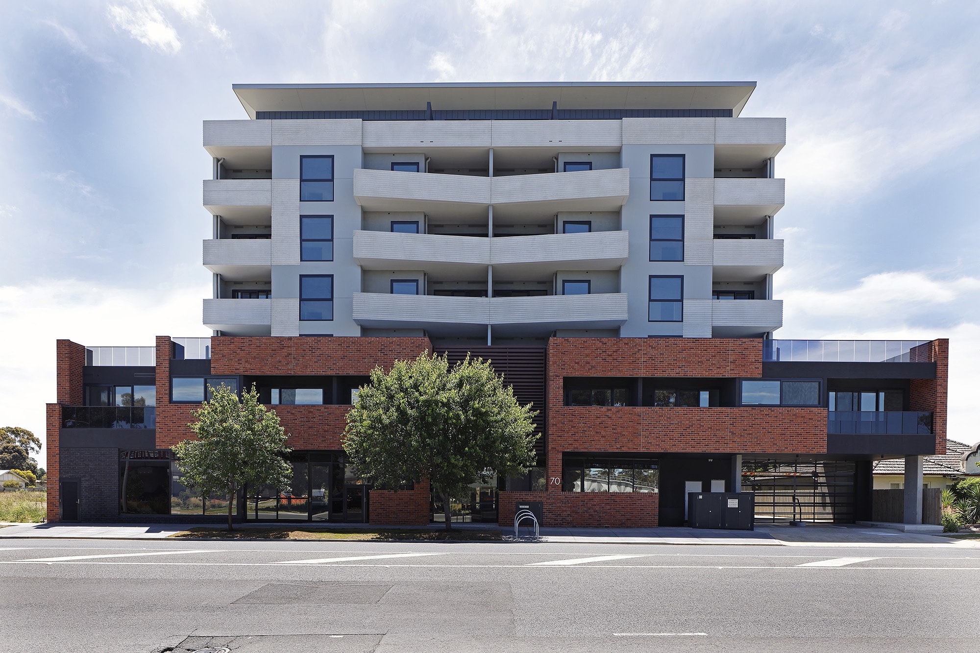 A modern multi-story apartment building with a red brick lower façade and light gray upper levels, large balconies, and trees in front, viewed from across a wide street.