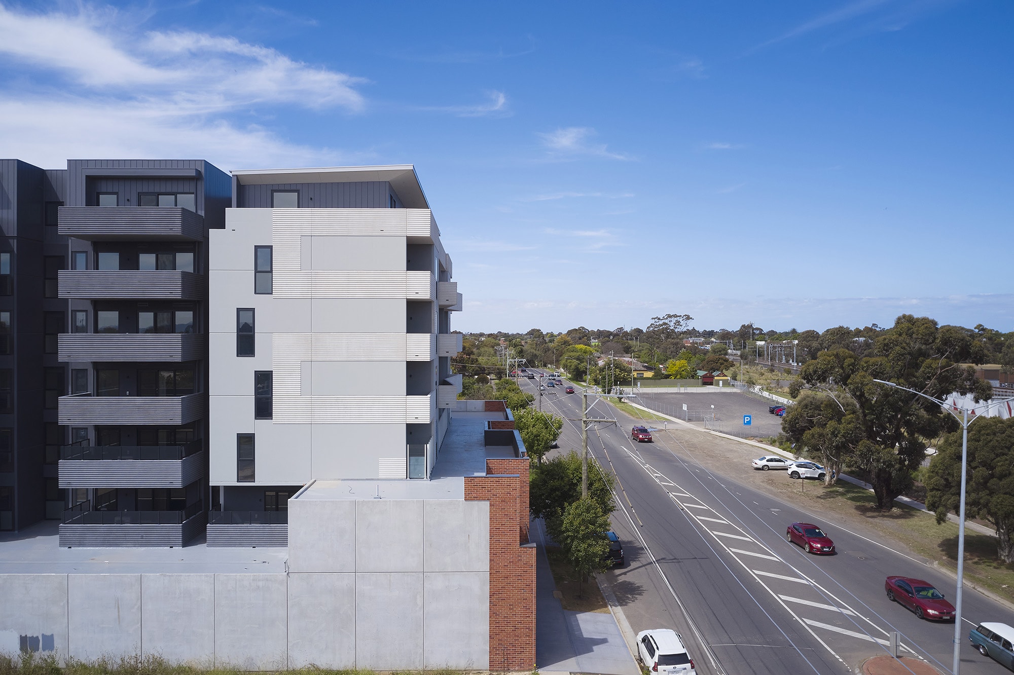 A modern multi-story apartment building stands beside a wide road with several cars, trees, and open spaces under a blue sky with scattered clouds.
