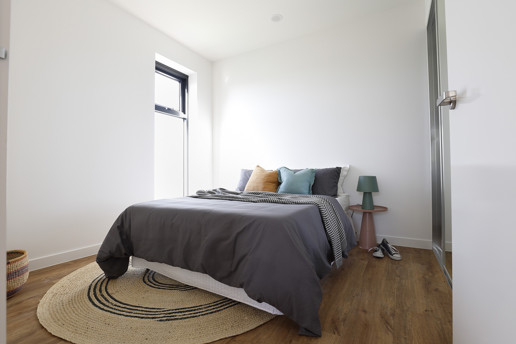 A modern, minimal bedroom with a double bed covered in gray bedding, several throw pillows, a round rug, a small bedside table with a green lamp, and shoes on a wooden floor near the bed.