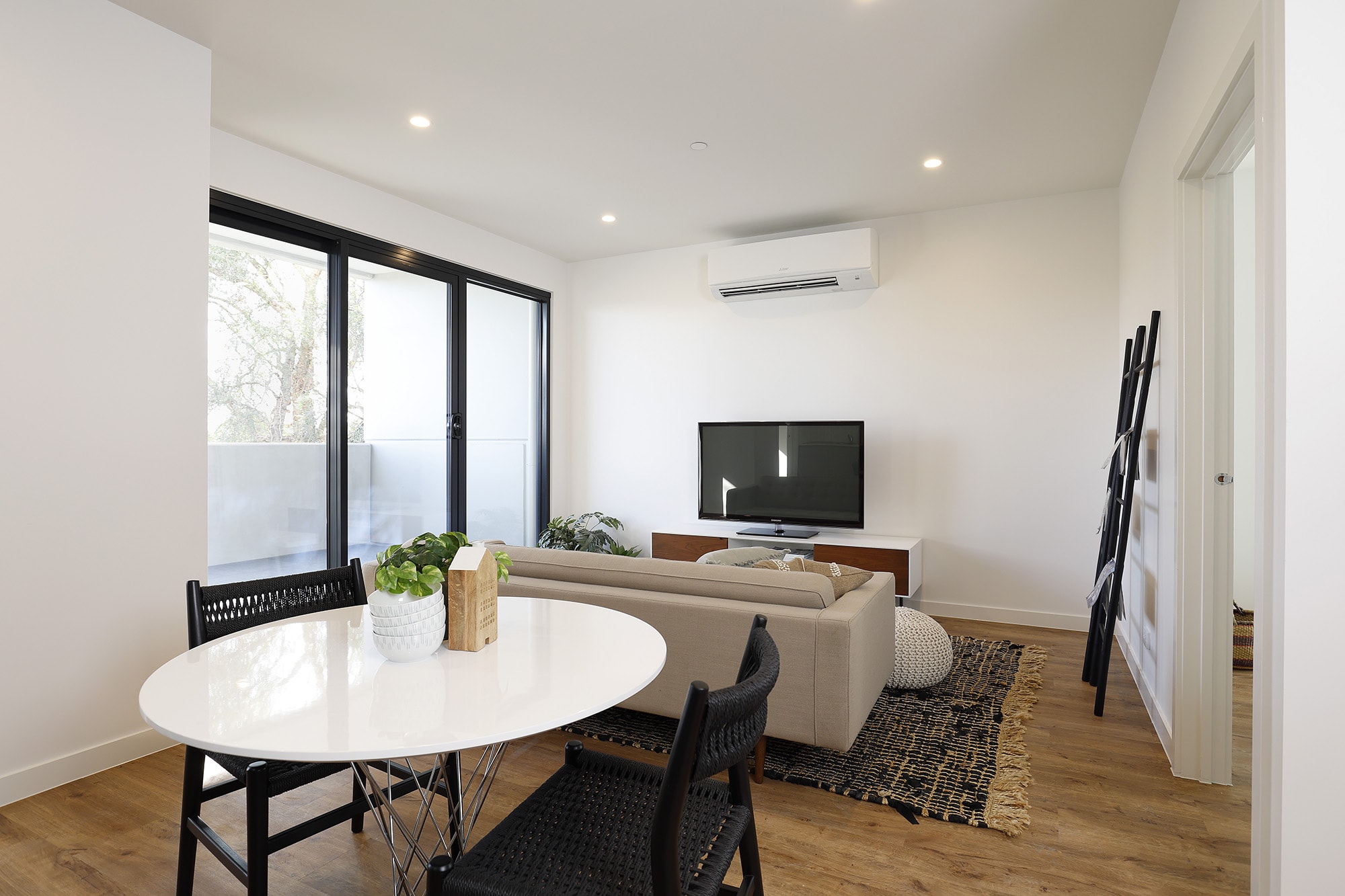 Modern living room with a beige sofa facing a TV on a wooden stand, round white dining table with black chairs, sliding glass doors, and wooden flooring. A ladder-style shelf and indoor plant add decorative touches.