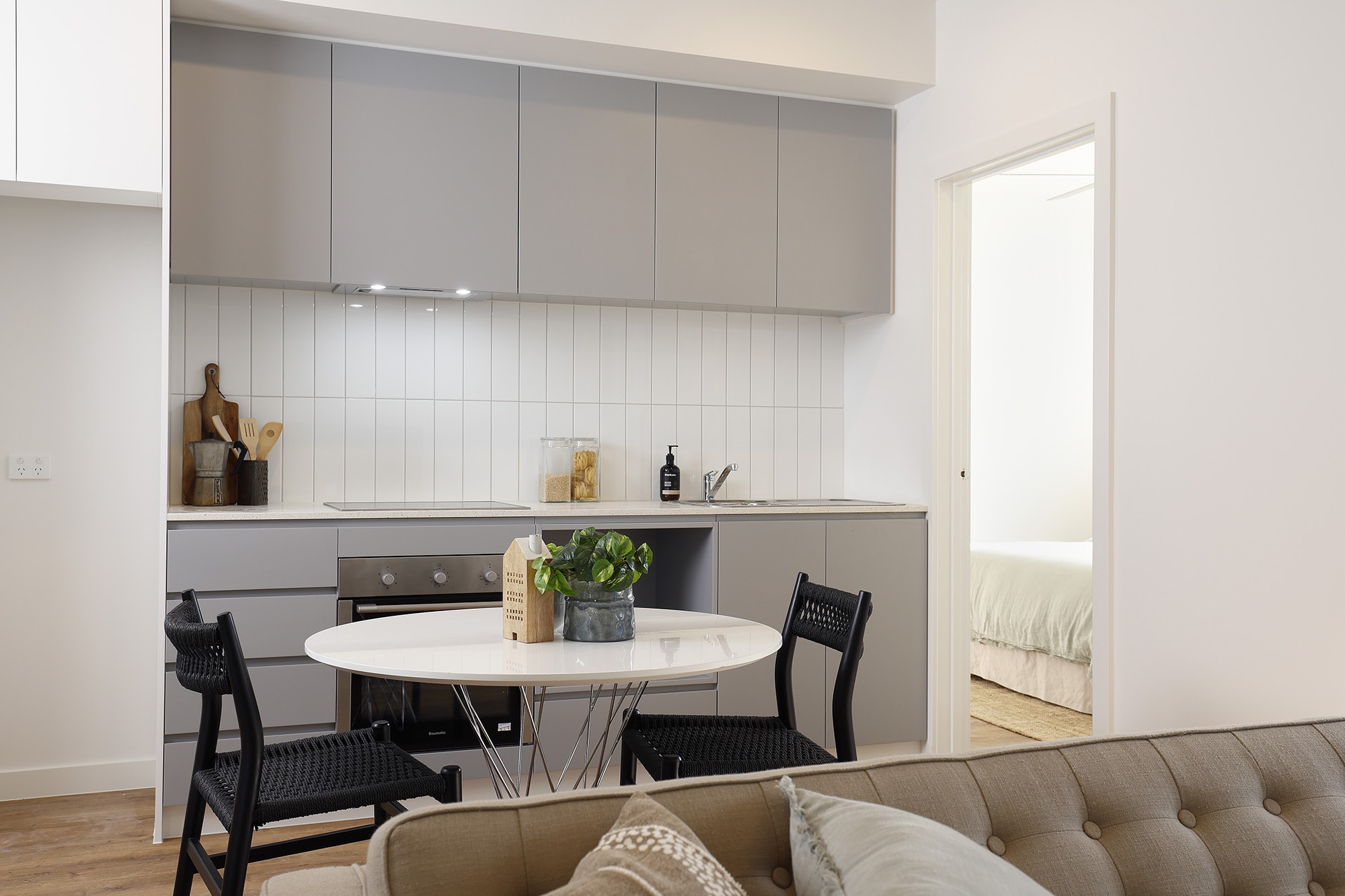 Modern kitchen with grey cabinets and a small round dining table set for two with black chairs. A plant centerpiece is on the table. In the background, a doorway leads to a bedroom with a neatly made bed.