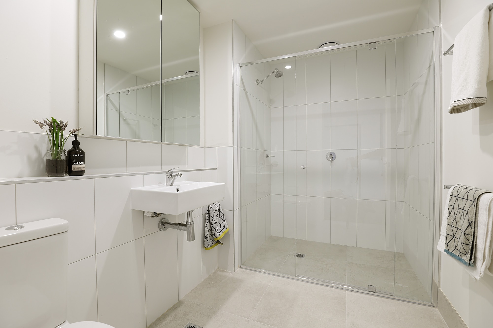 Modern bathroom with a glass-enclosed shower, wall-mounted sink, toilet, large mirror, towels hanging on racks, and minimal decor including a small plant and soap dispenser on the sink.