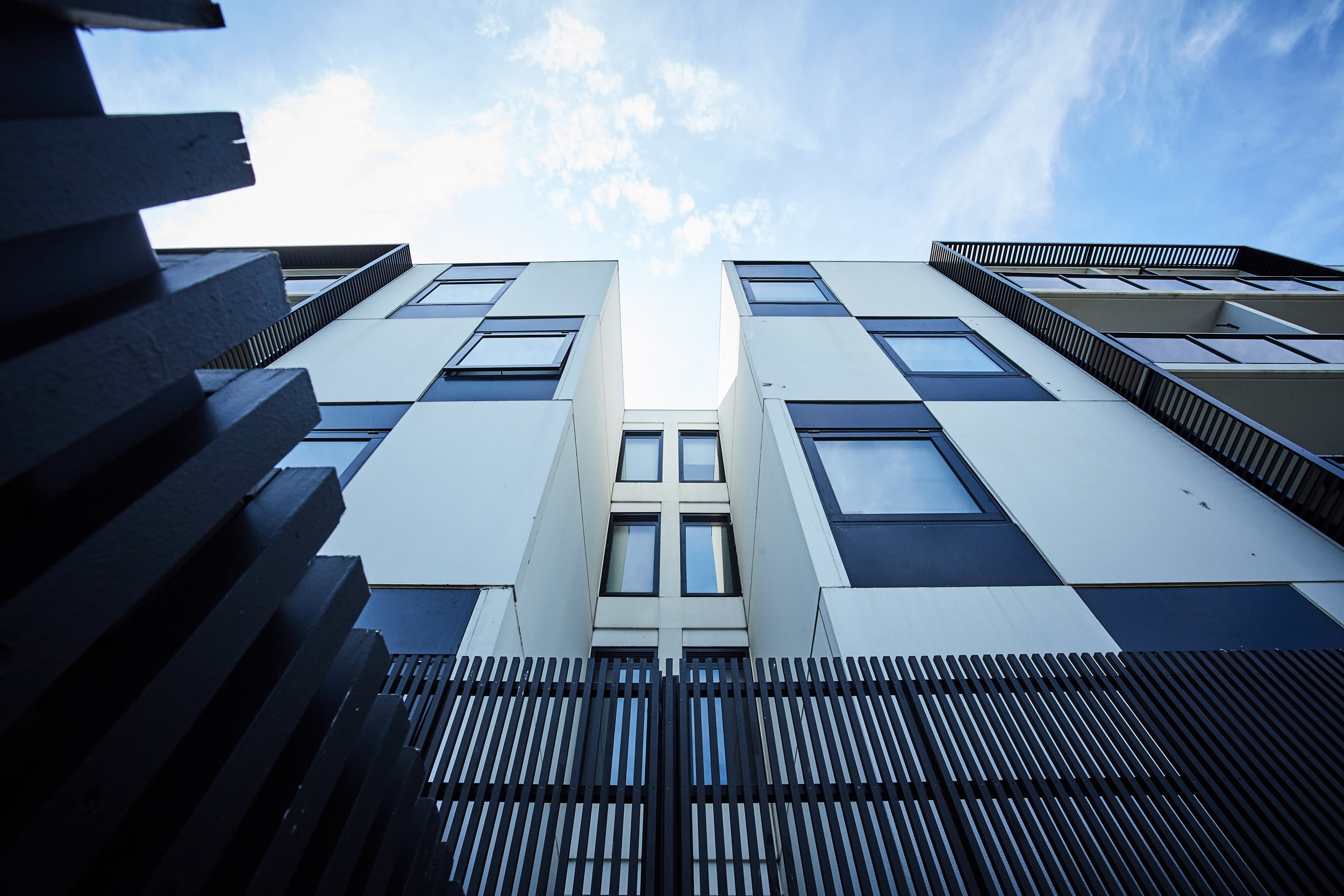 Low-angle view of a modern apartment building with large windows, black and white geometric facade, and a metal fence in the foreground against a blue sky with light clouds.