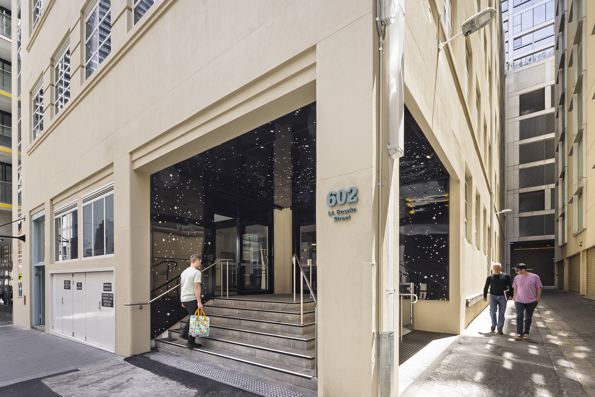 A man with a colorful bag walks up the stairs to the entrance of a beige building at 602 St. Kilda Road, while two people walk past on the sidewalk. Sunlight casts shadows on the building and pavement.