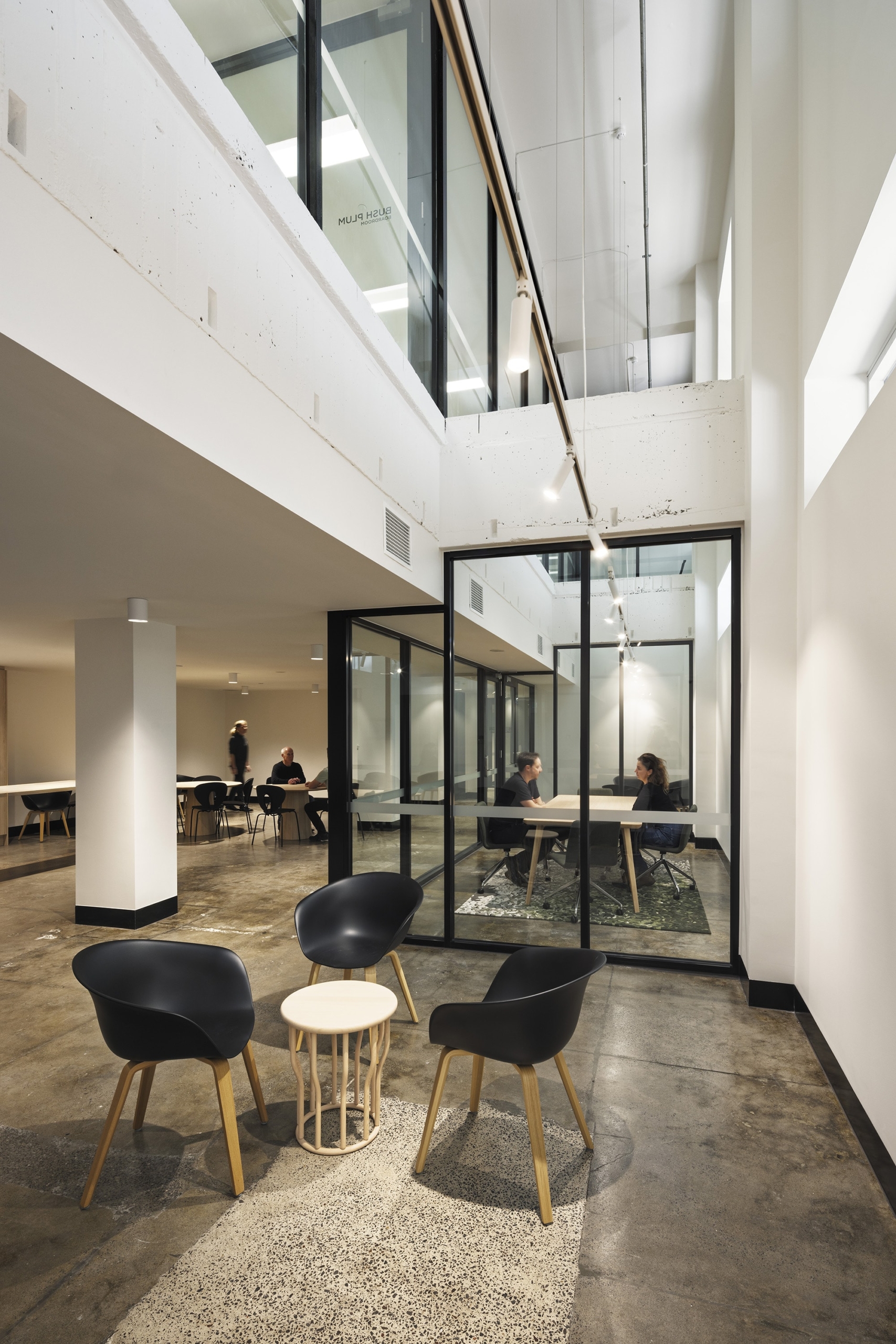 Modern office space with high ceilings, black chairs around a small round table, glass-walled meeting rooms, and people working at tables in the background. The area is well-lit with a clean, minimalist design.