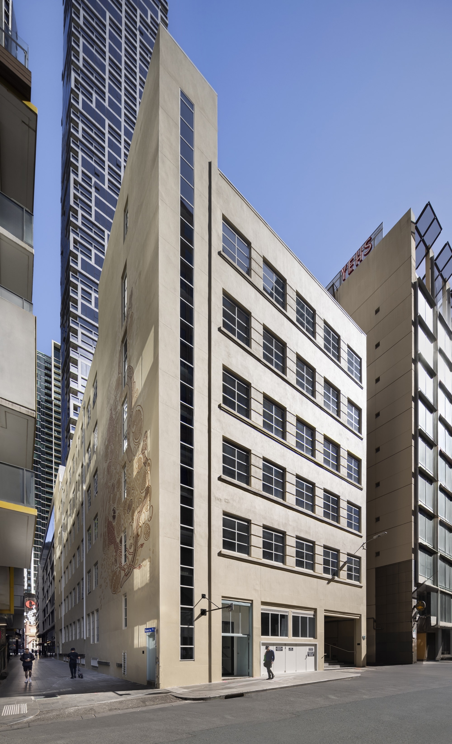 A beige mid-rise building with large rectangular windows and a vertical glass column stands on a city street, with a mural visible on one wall. Tall modern skyscrapers and buildings surround it under a clear blue sky.