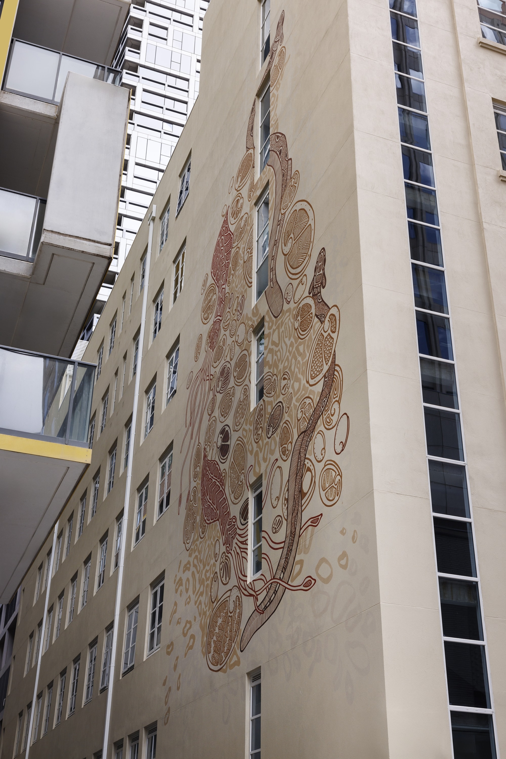 A large mural of abstract shapes, fruit slices, and plant elements decorates the side of a tall beige building with many windows in an urban setting.
