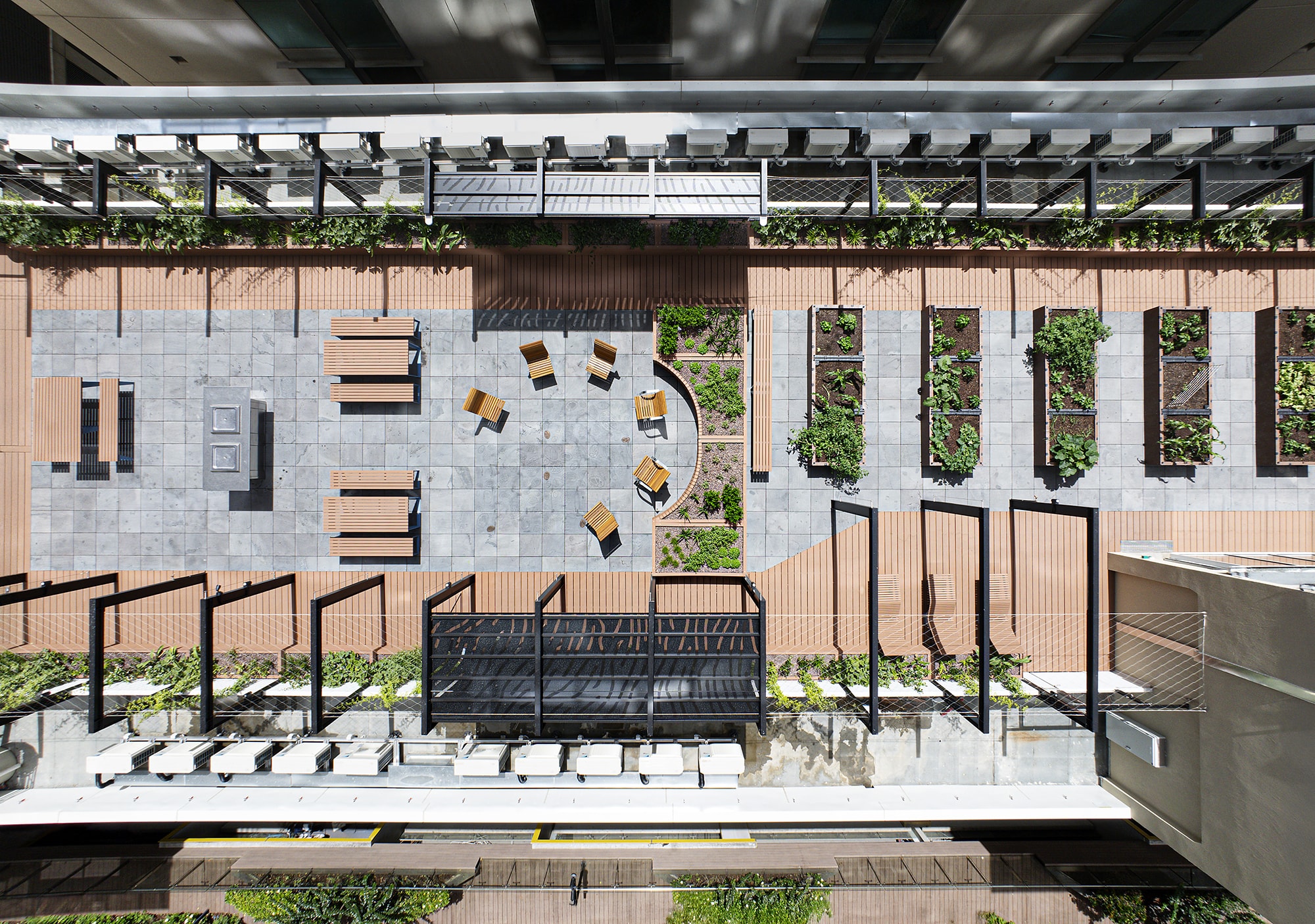 Aerial view of a modern rooftop terrace with benches, tables, planter boxes with greenery, and seating arranged in a semicircle on a tiled floor. The area is bordered by lined walkways and surrounded by railings.