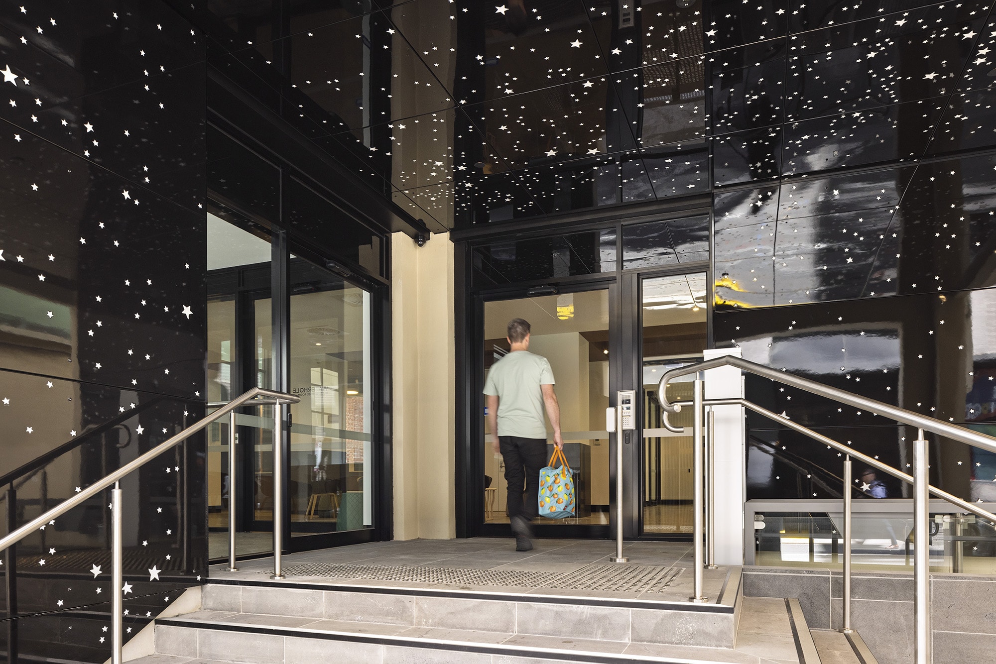 A man carrying a colorful bag walks into a building with shiny black walls decorated with small white star patterns, seen from the outside entrance steps.