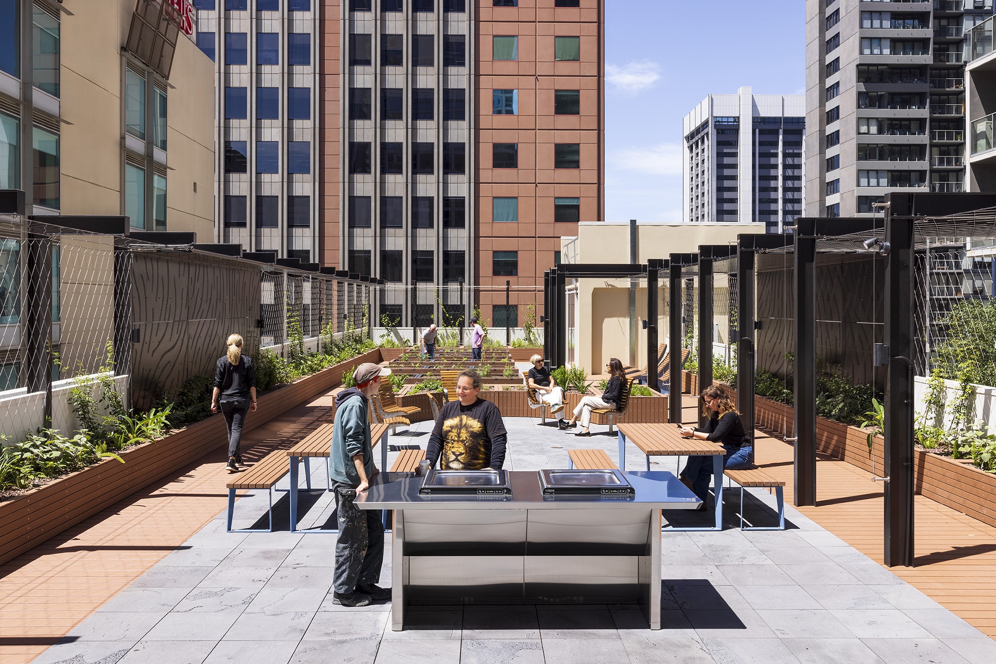 People relax and socialize on a modern rooftop terrace in a city, featuring benches, plants, pergolas, and a stainless steel barbecue area, surrounded by tall office buildings under a partly cloudy sky.