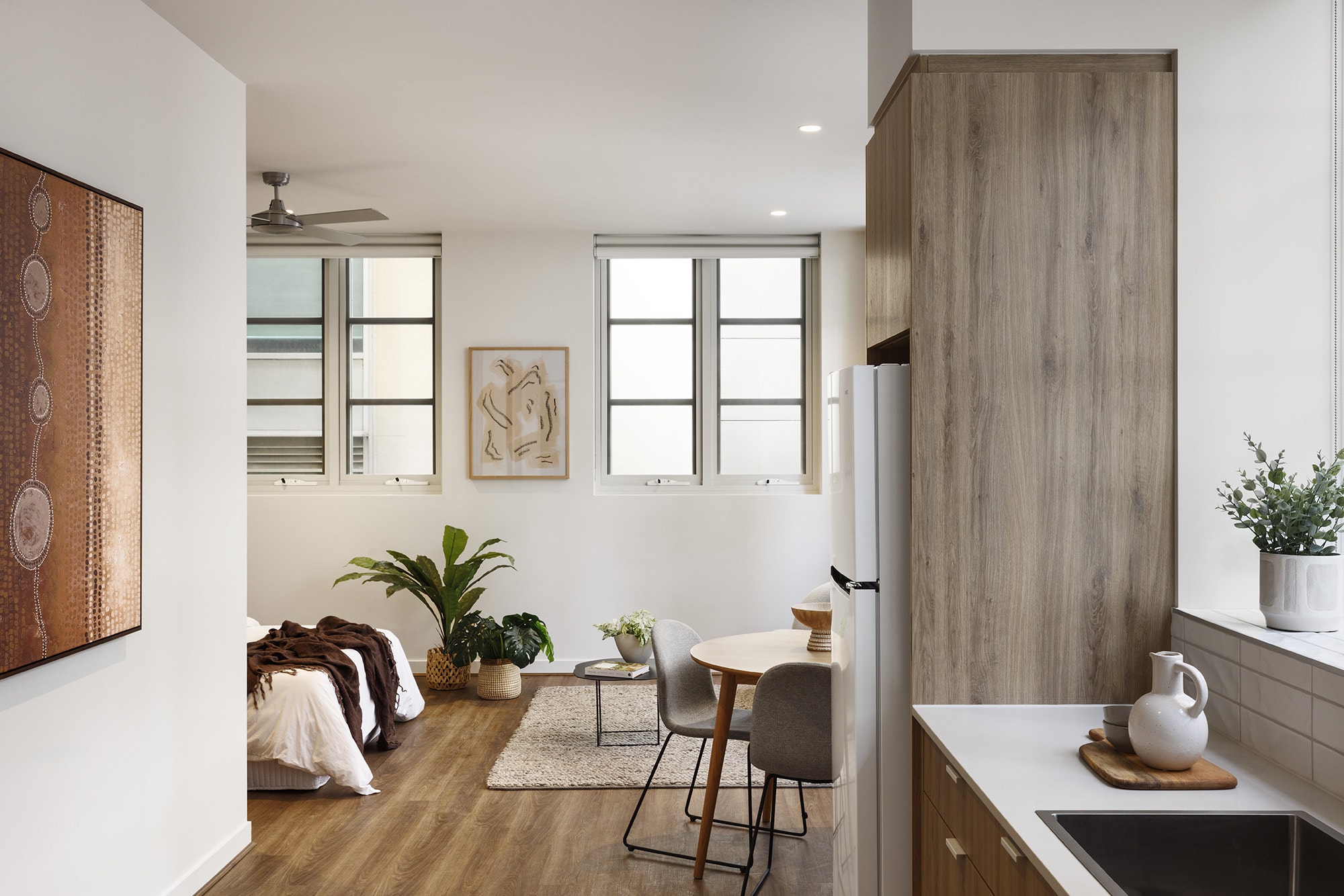 A modern studio apartment with a bed, small dining table, chairs, plants, and wall art. The kitchen area has wood cabinets, a white countertop, and a potted plant. Large windows let in natural light.