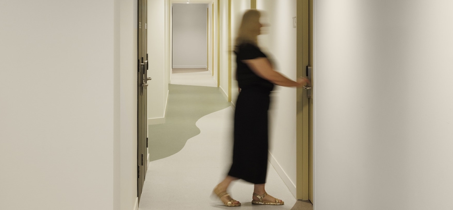 A blurred person in black clothing walks down a modern hallway with white walls and multiple closed doors, about to open one of the doors.