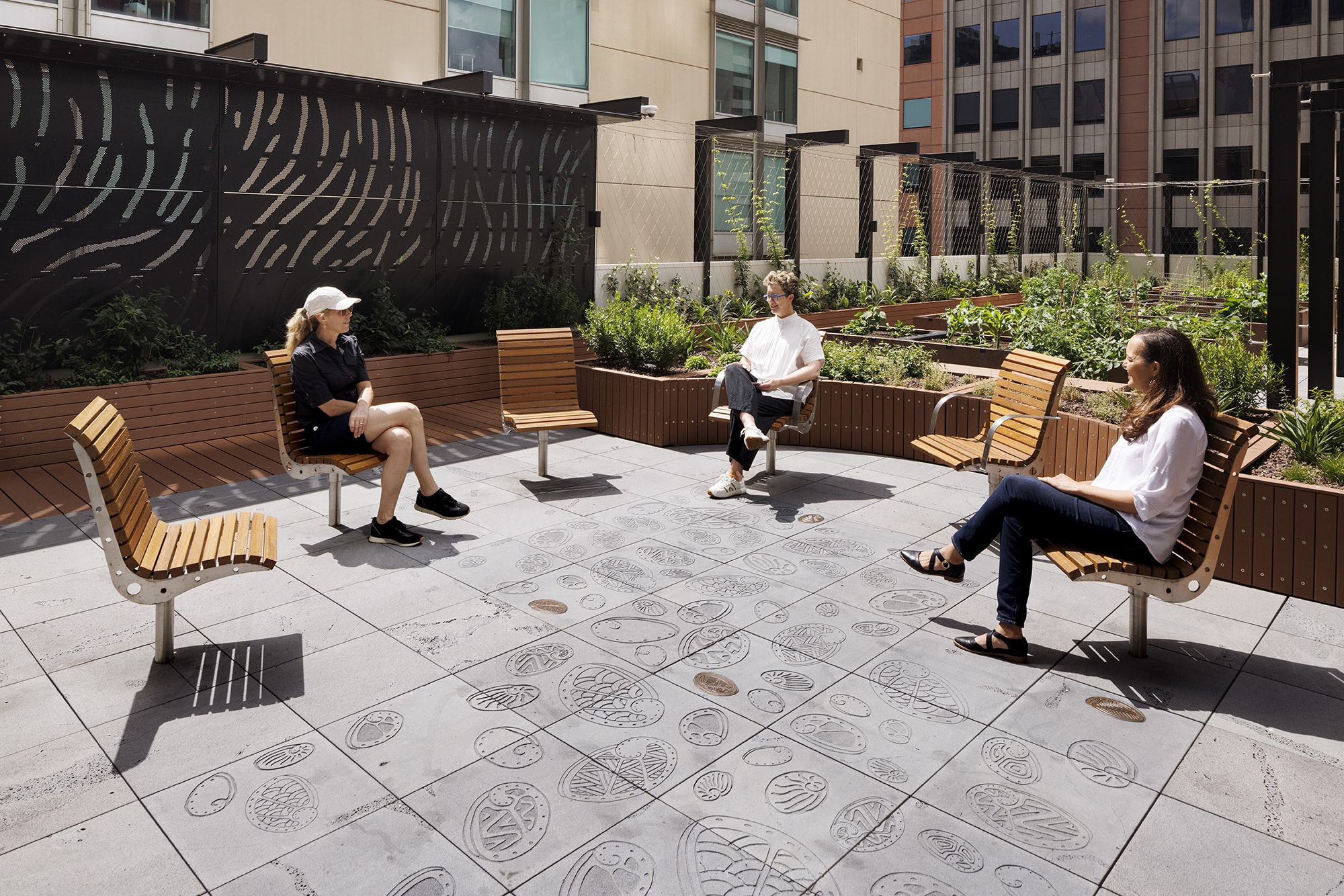 Three people sit on wooden benches spaced apart in an outdoor urban garden with raised planters, engraved paving stones, and surrounding buildings in the background on a sunny day.