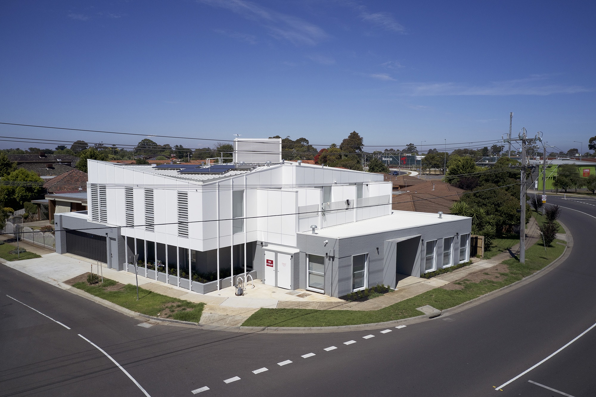 A modern white building with flat roofs and large windows sits on a street corner with surrounding asphalt roads, sidewalks, and residential houses in the background under a clear blue sky.