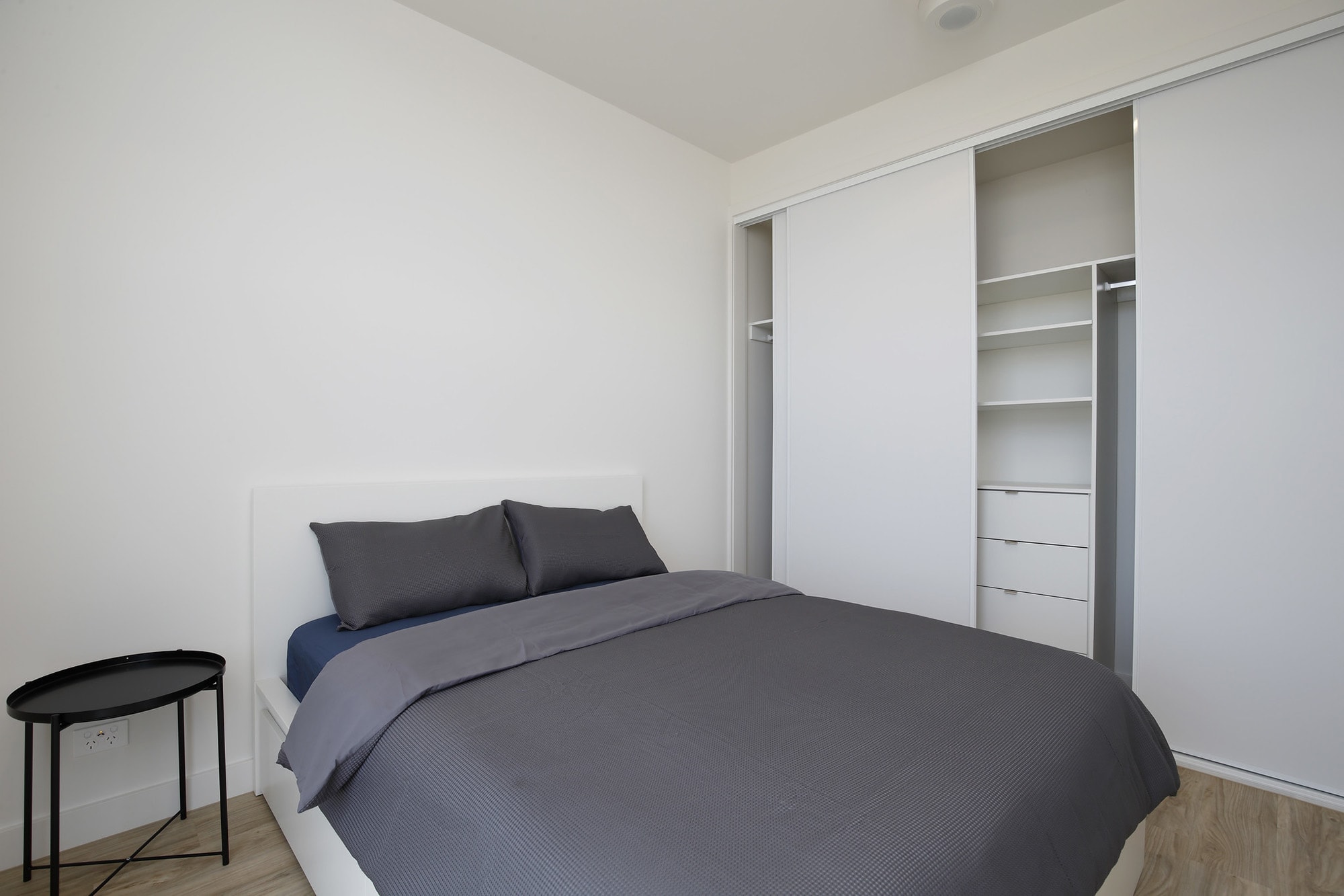 A minimal bedroom with a neatly made bed featuring gray bedding, a small round black side table, and an open closet with sliding white doors and built-in shelves. The walls and ceiling are white.
