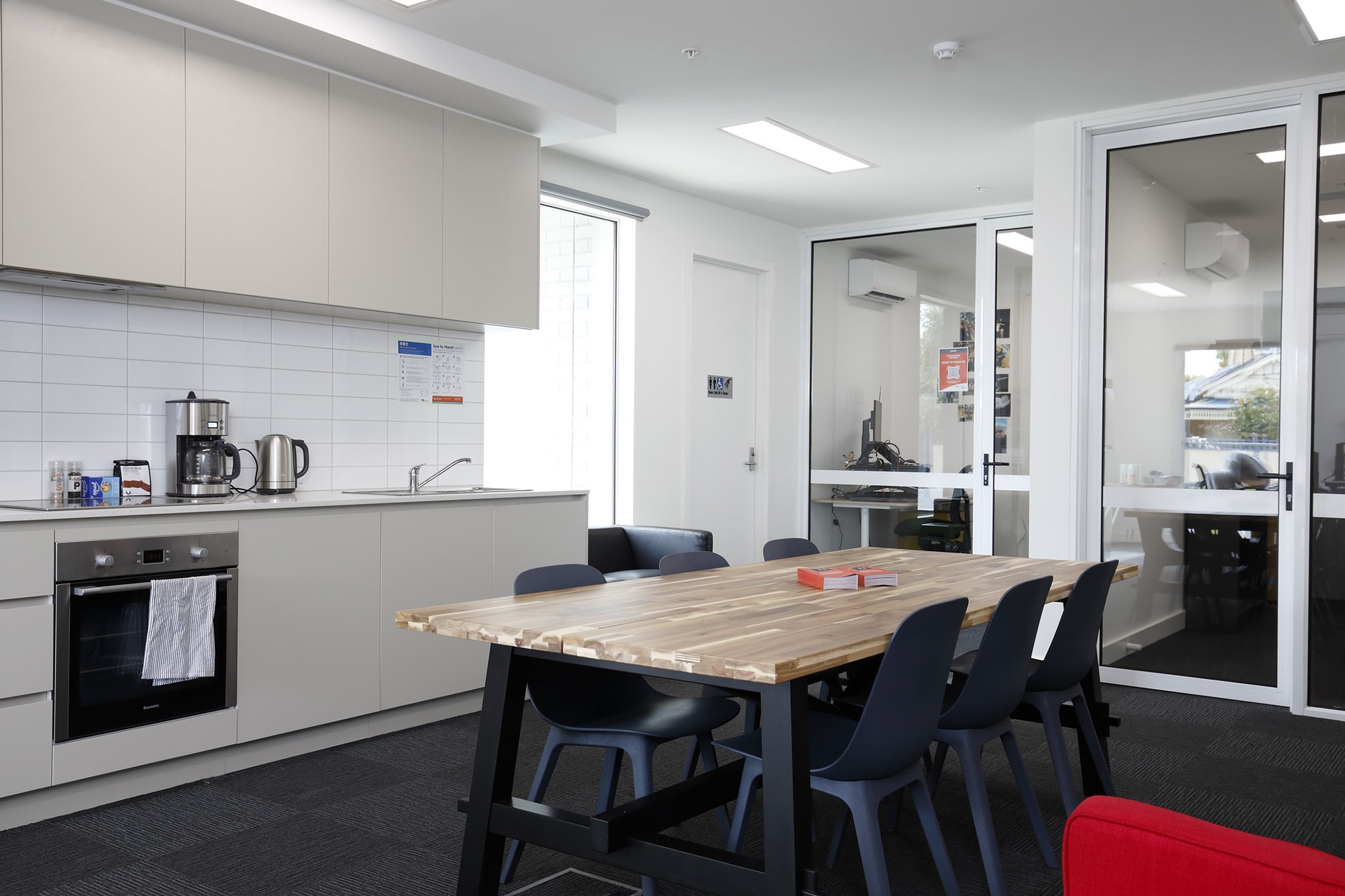 Modern kitchen and dining area with a wooden table, black chairs, beige cabinets, appliances, and a glass door leading to an office. Bright, clean, and minimalistic space with natural light.