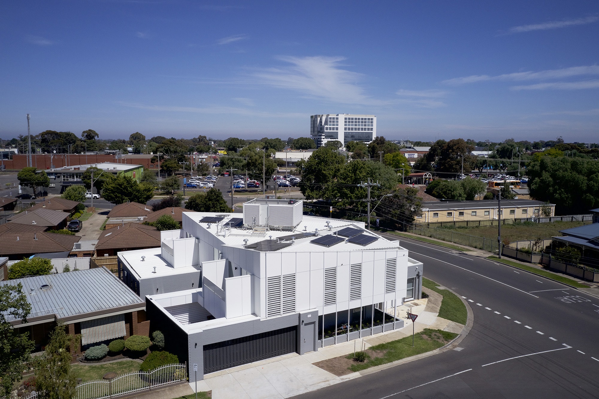 A modern white building with solar panels on the roof sits on a corner lot, surrounded by suburban houses and greenery. A multi-story office building and parking lot are visible in the background under a clear blue sky.
