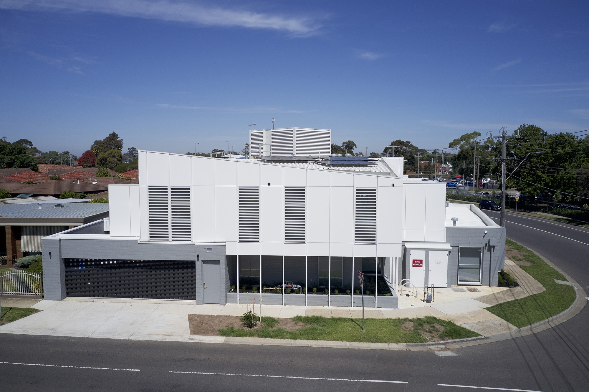 A modern, white, two-story building with large vertical windows and vents, located on a corner lot near residential houses and a street intersection, under a clear blue sky.