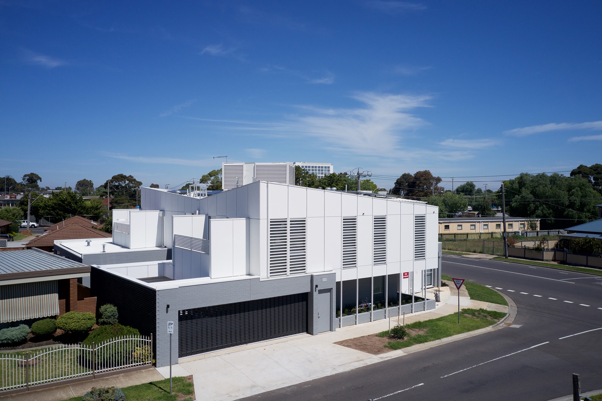 A modern, white building with large windows and louvered panels sits on a street corner under a clear blue sky, surrounded by small shrubs and nearby houses.