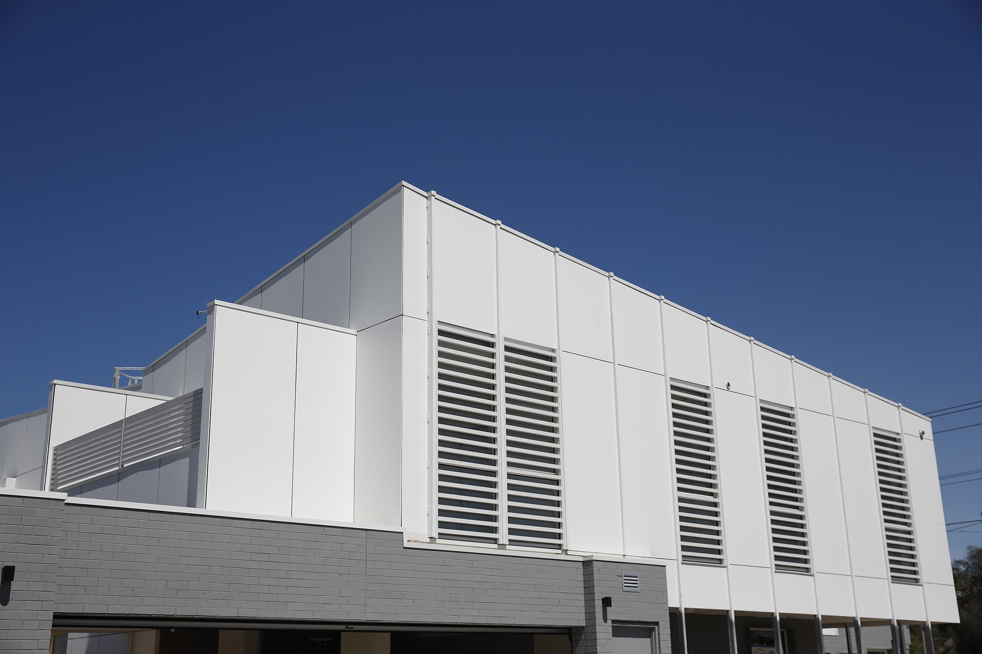 A modern building with white paneled walls and vertical slats, featuring geometric lines and a gray base, set against a clear blue sky.