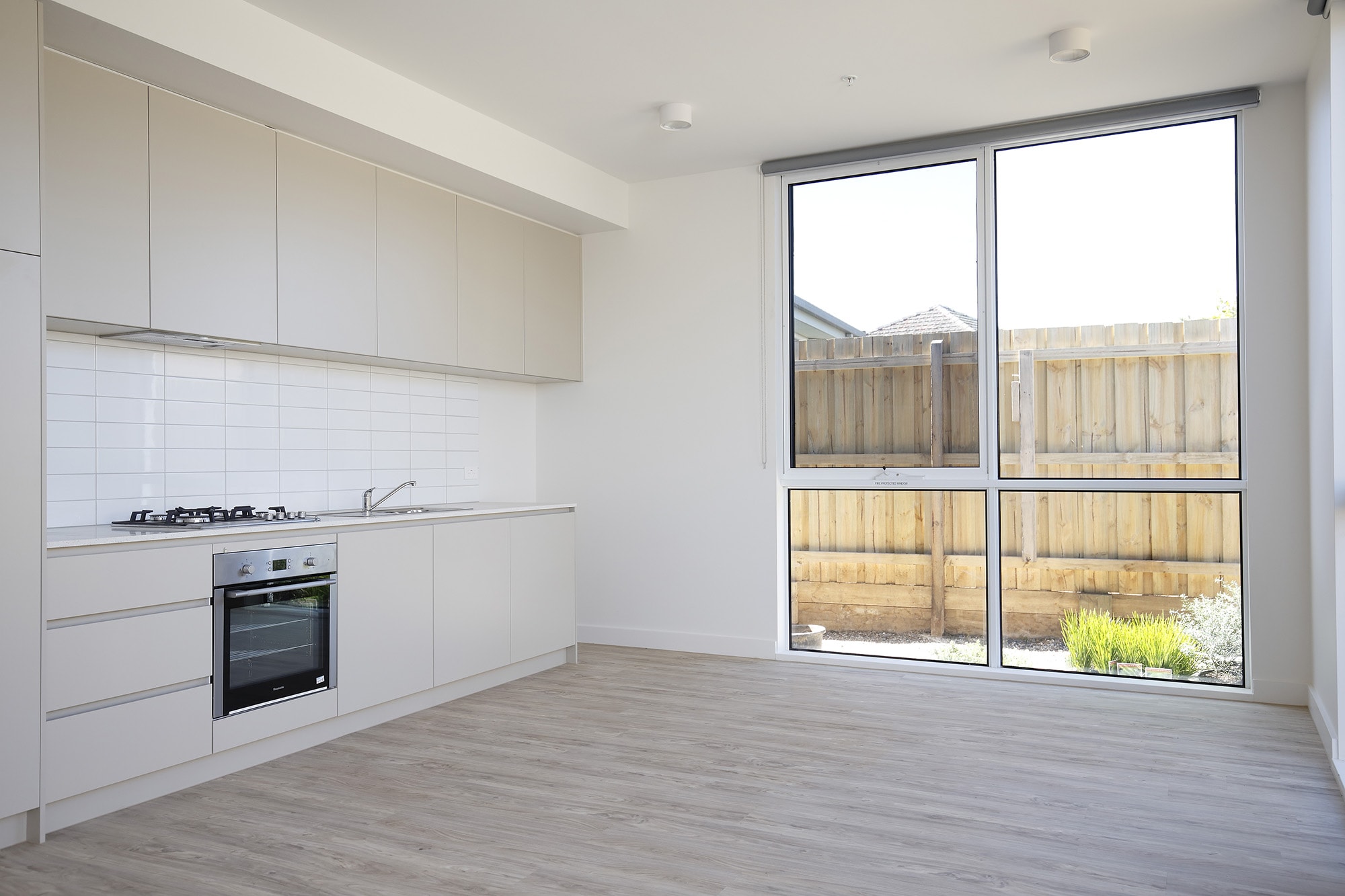 Modern, minimal kitchen with light cabinets, oven, gas stove, and sink. Large windows let in natural light and overlook a fenced backyard with plants. The floor is light wood, and the overall space is bright and clean.