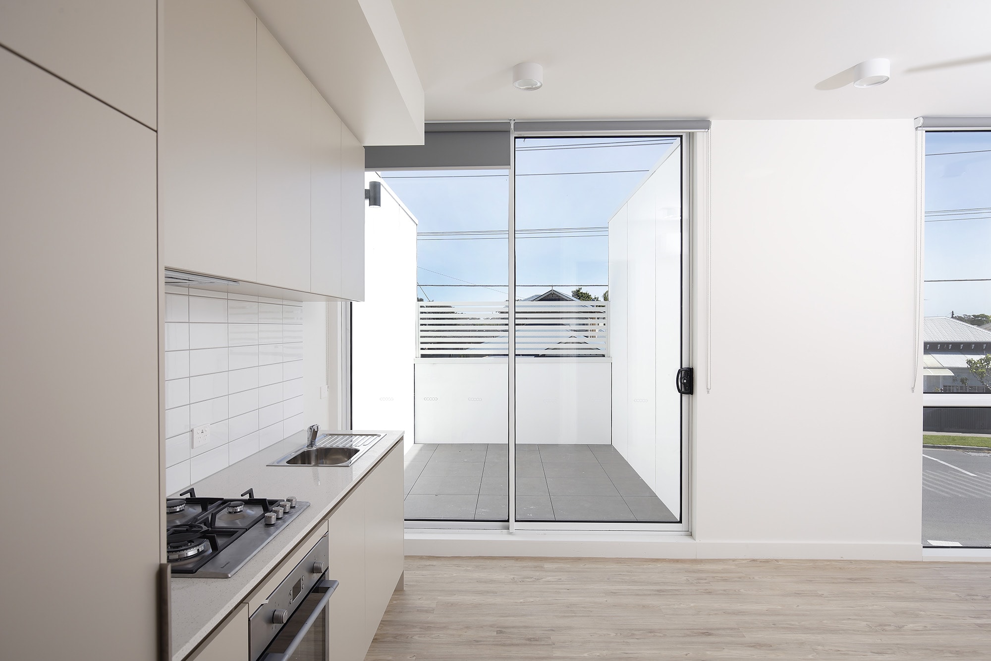 Modern kitchen with light-colored cabinets, a gas stove, and a sink, next to large glass sliding doors that open to a small balcony with white walls and a view of neighboring buildings.