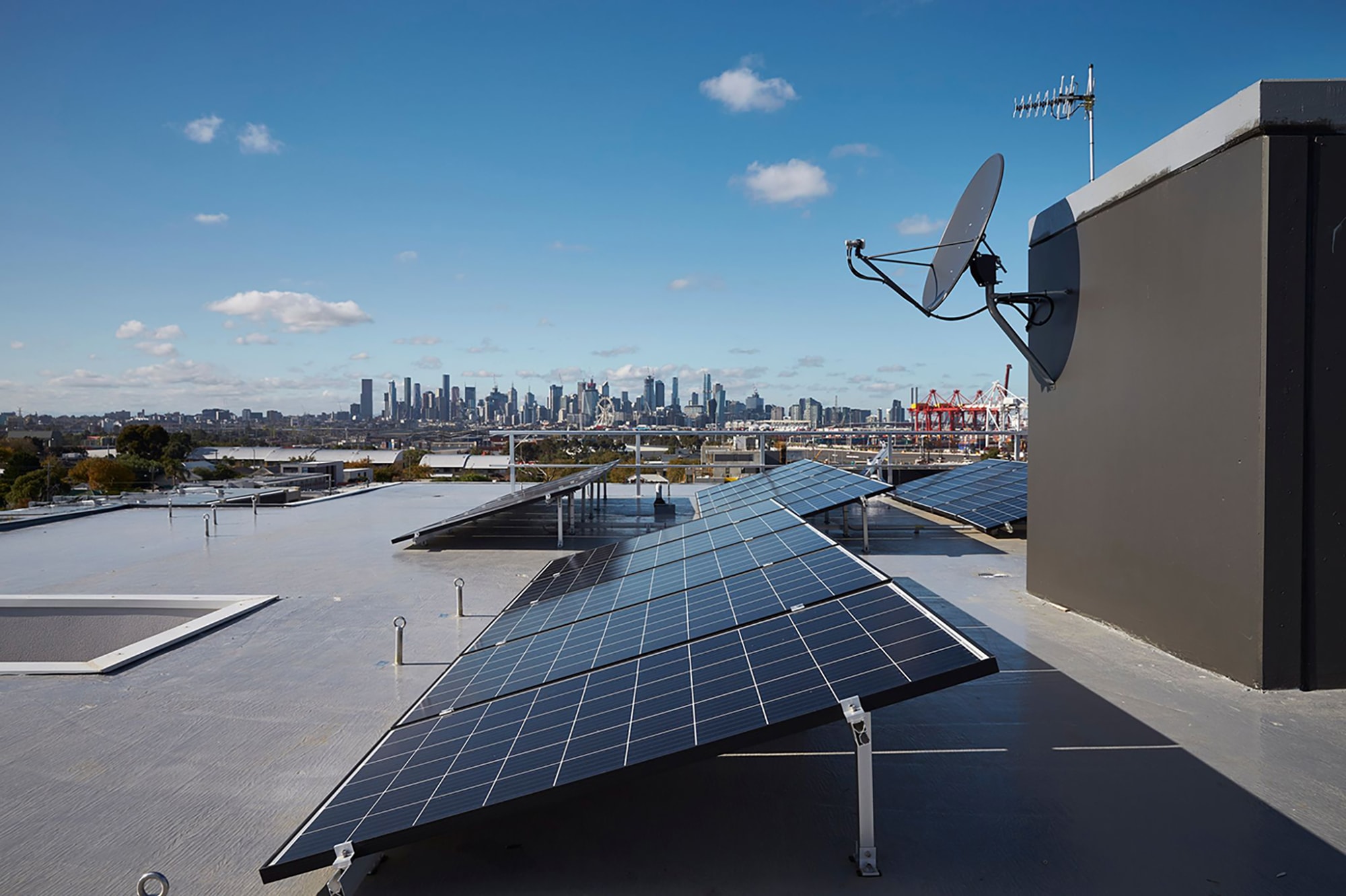 Several solar panels are installed on a flat rooftop with a satellite dish nearby; a city skyline and blue sky with scattered clouds are visible in the background.