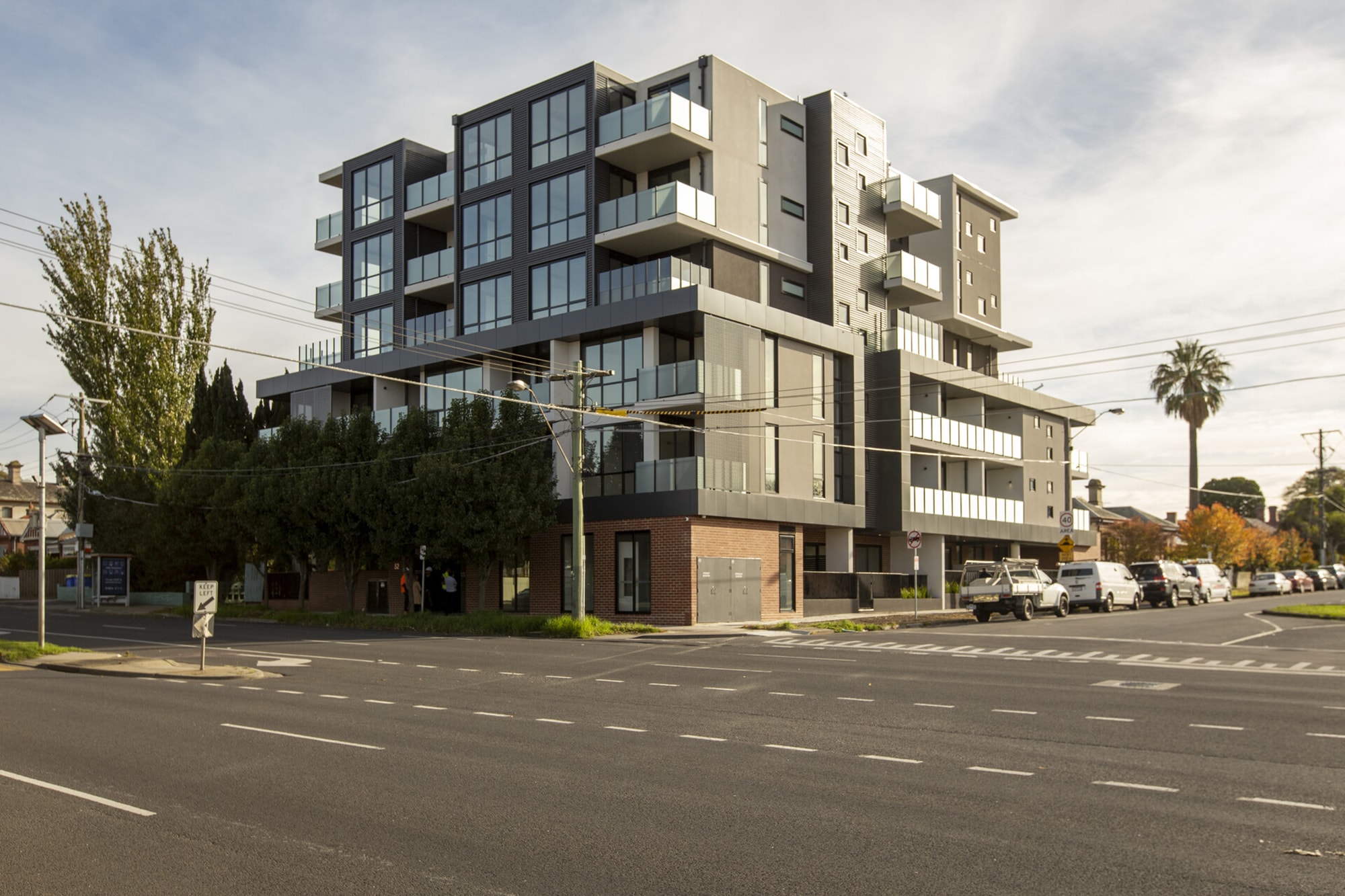 A modern multi-story apartment building with large windows and balconies sits on a street corner, surrounded by trees, parked cars, and intersecting roads under a partly cloudy sky.