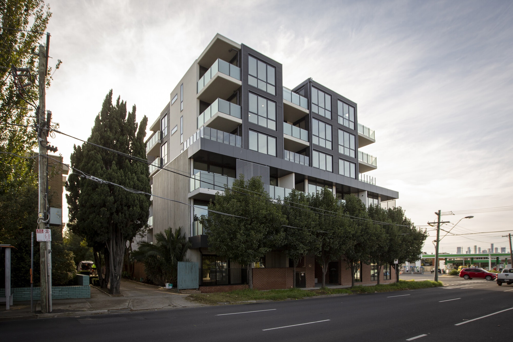 A modern, multi-story apartment building with large windows and balconies, surrounded by trees, situated on a street corner with cars and city skyline visible in the distance.