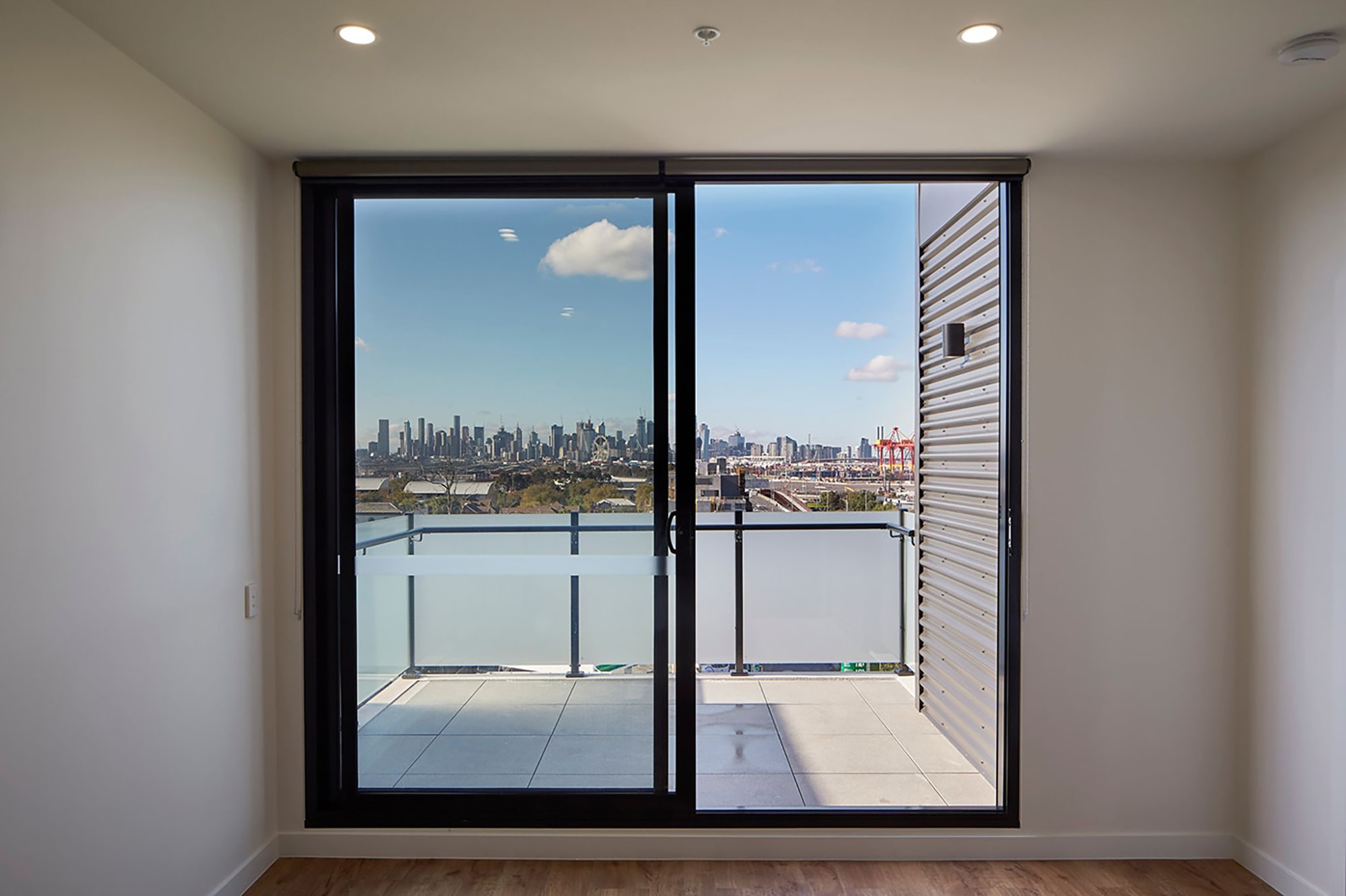 A glass sliding door opens to a balcony with a clear view of a city skyline under a blue sky with a few clouds. The room has wooden flooring and white walls.
