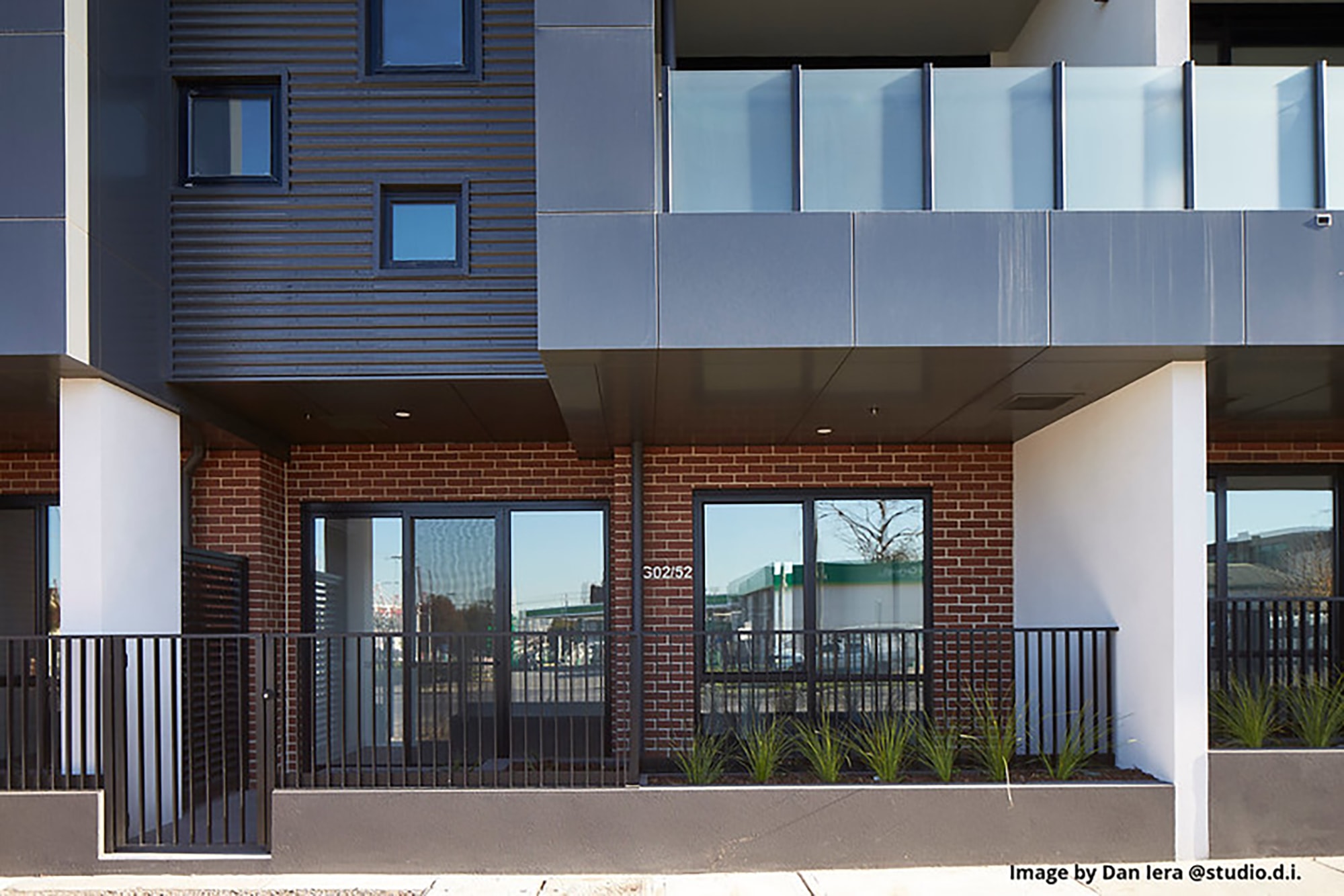 Street view of a modern apartment building with large windows, a brick and metal exterior, black railing, and a row of small green plants in front. The address 302/32 is visible near the entrance.