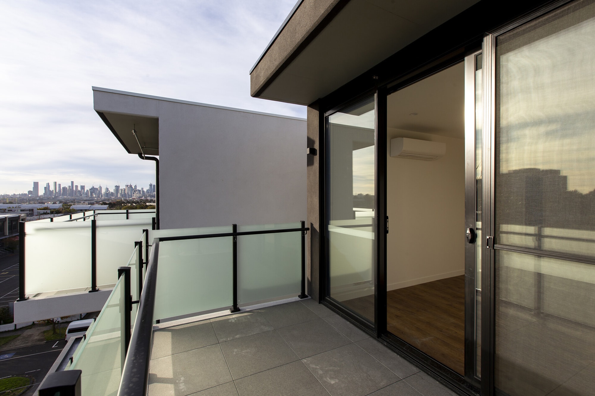 Modern apartment balcony with frosted glass railings, overlooking a city skyline in the distance. Sliding glass doors open to an empty room with wooden floors. The sky is partly cloudy.