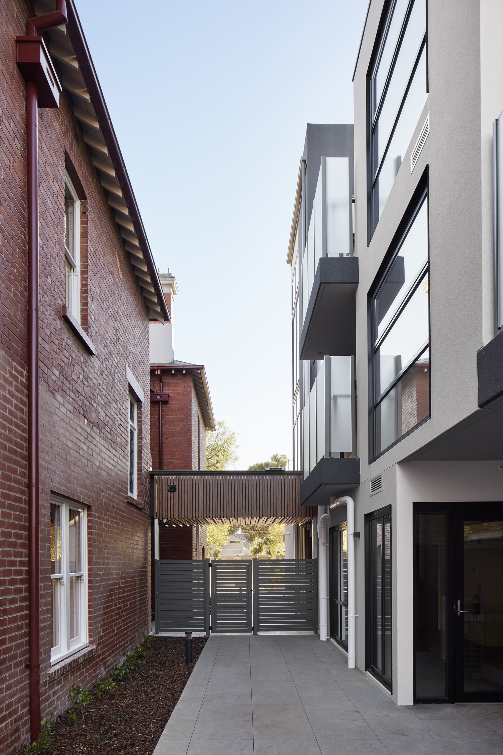 A narrow walkway separates a traditional red brick building on the left from a modern, gray and white building on the right, with a metal gate and covered passageway visible ahead.