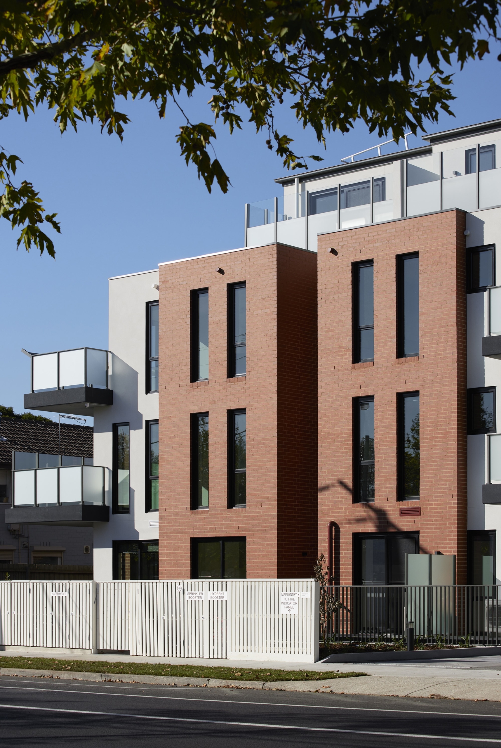 Modern apartment building with tall, narrow windows, red brick and white exterior walls, glass balconies, and a white fence in front. Tree branches partly frame the image, and the sky is clear and blue.
