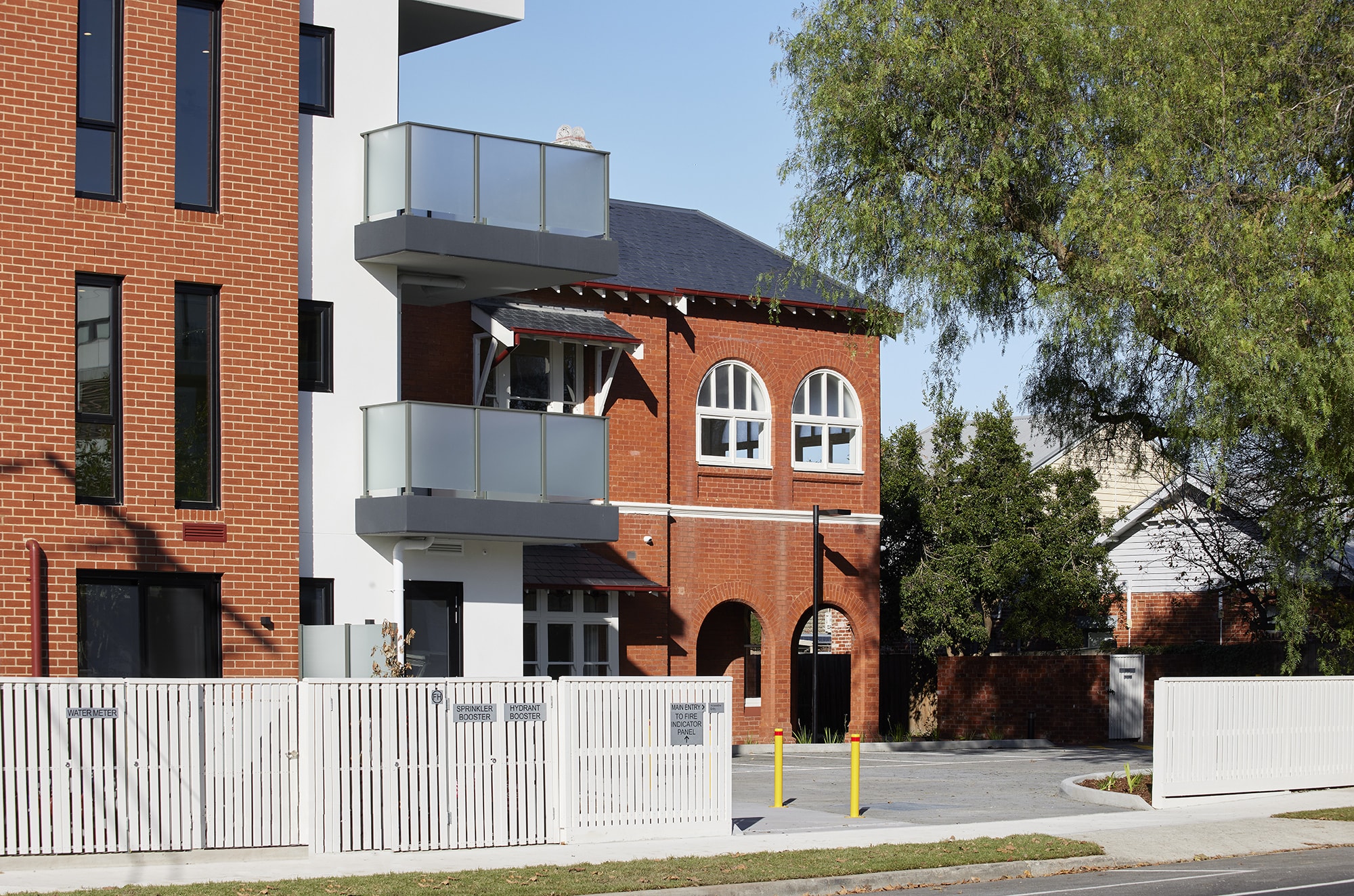 A modern apartment building with red brick and white sections, glass balconies, and a white fence stands next to a traditional red brick building with arched windows; trees line the street.