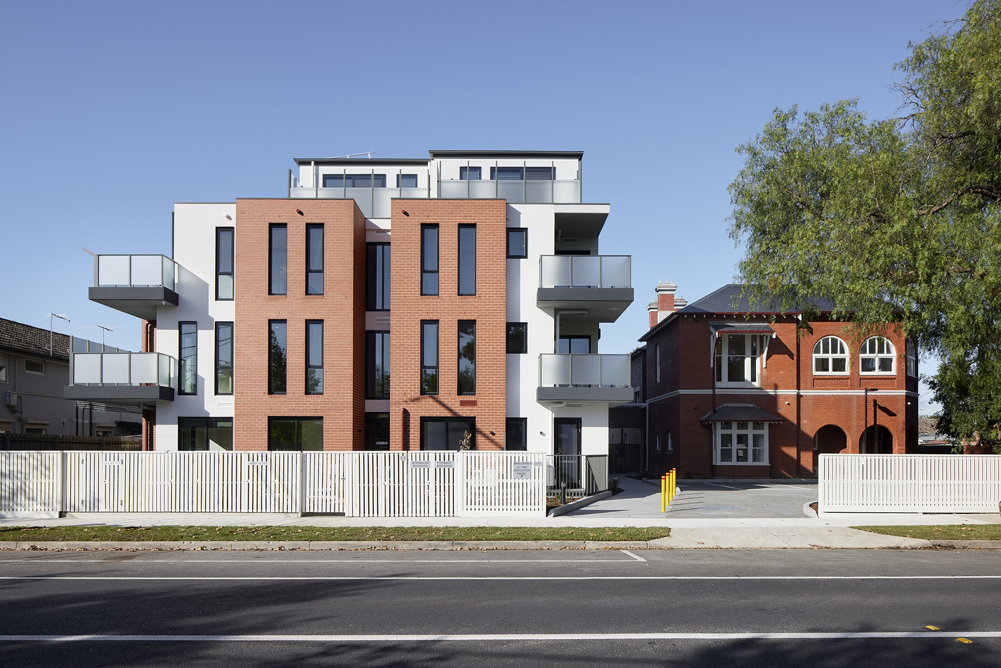 A modern apartment building with brick and white facades stands next to an older red brick house, both behind a white fence on a sunny day with clear blue sky.