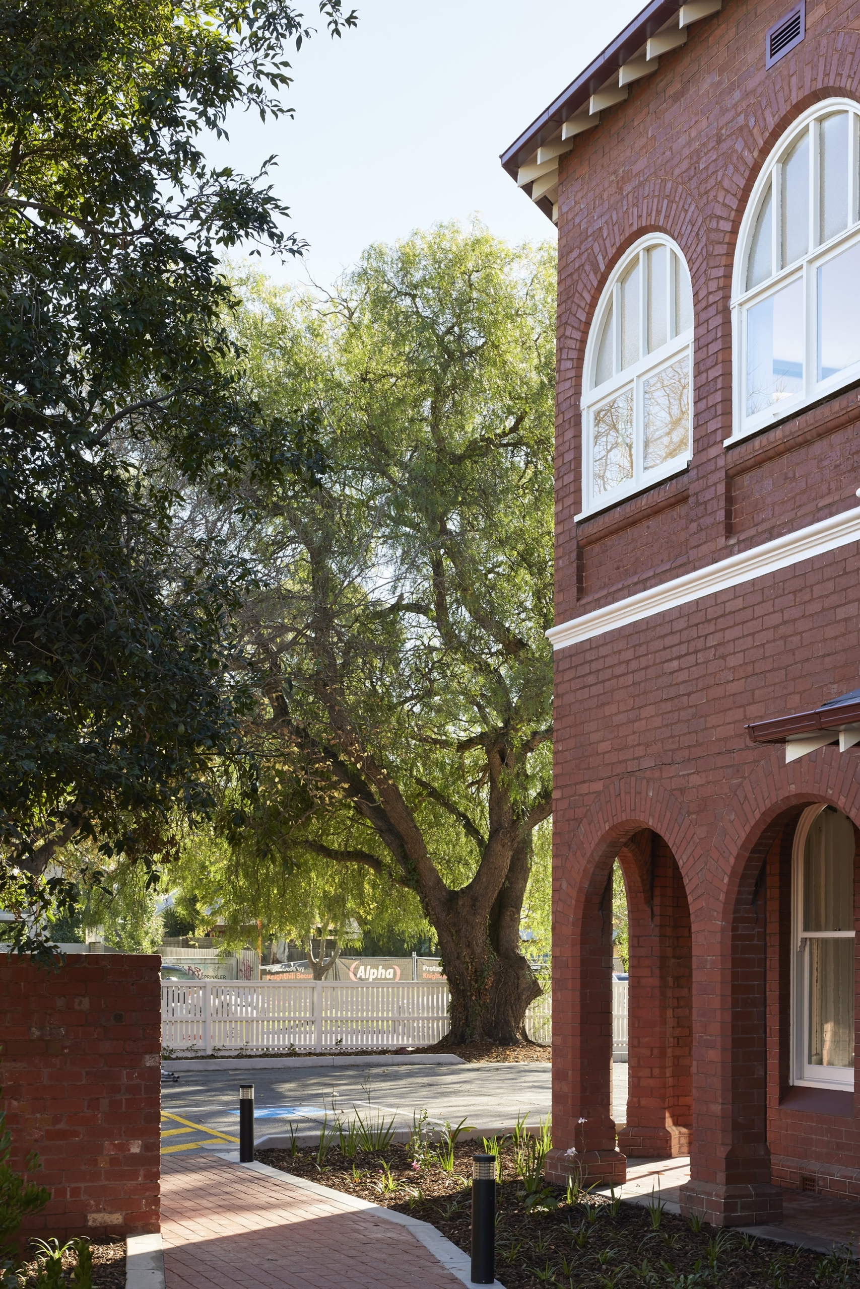 A large leafy tree stands near a red-brick building with arched windows and doorways on a sunny day. A paved path leads past the building, and a white picket fence is visible in the background.